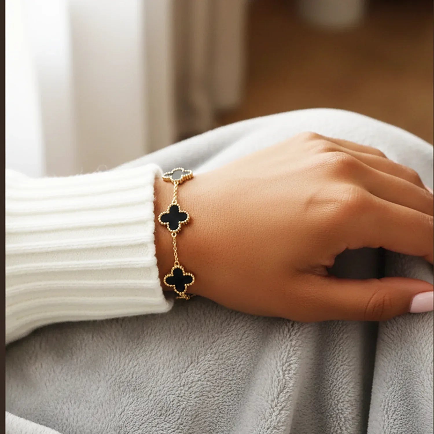 Hand wearing a gold bracelet with black clover charms on a neutral background