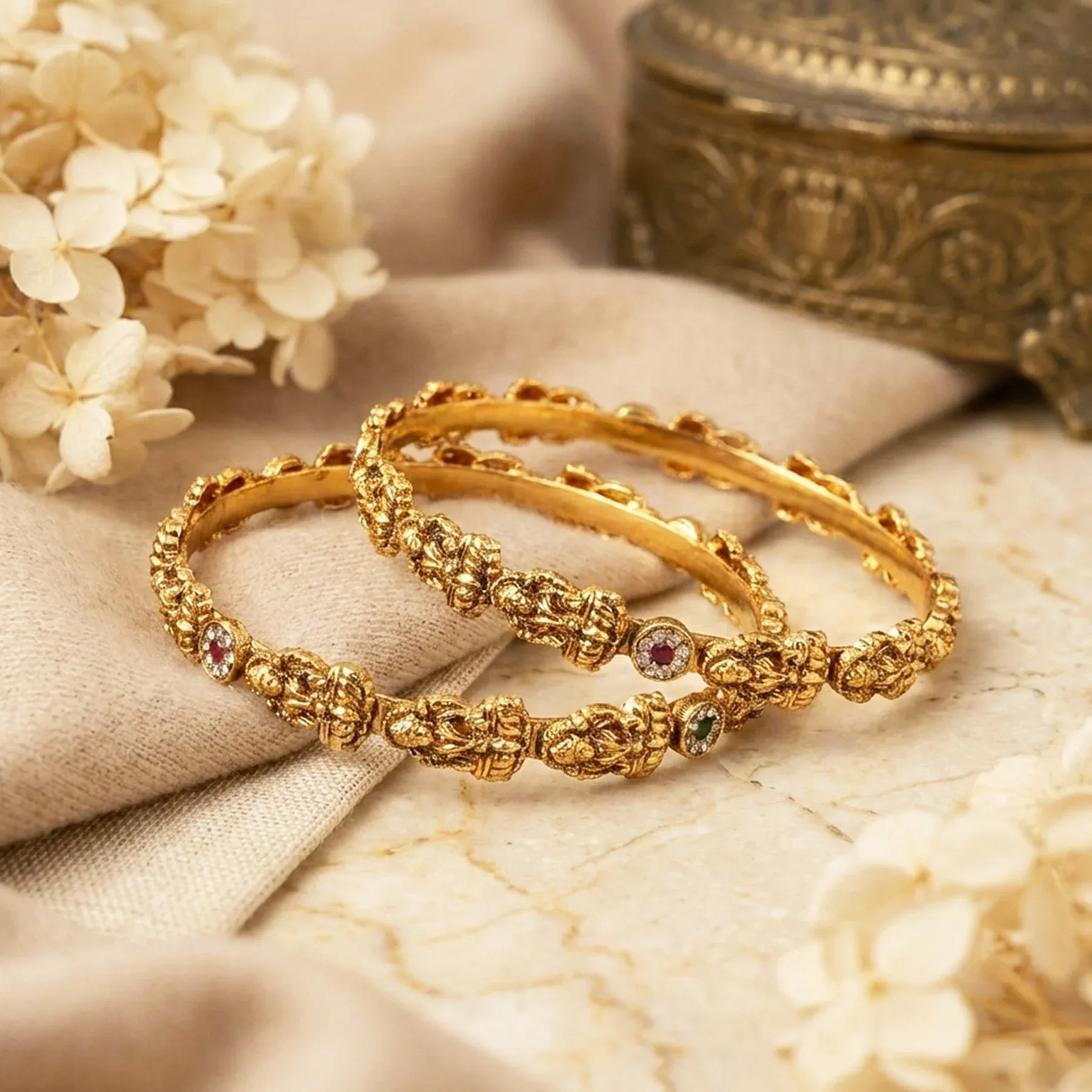 Two gold bangles with gemstones on a beige fabric background