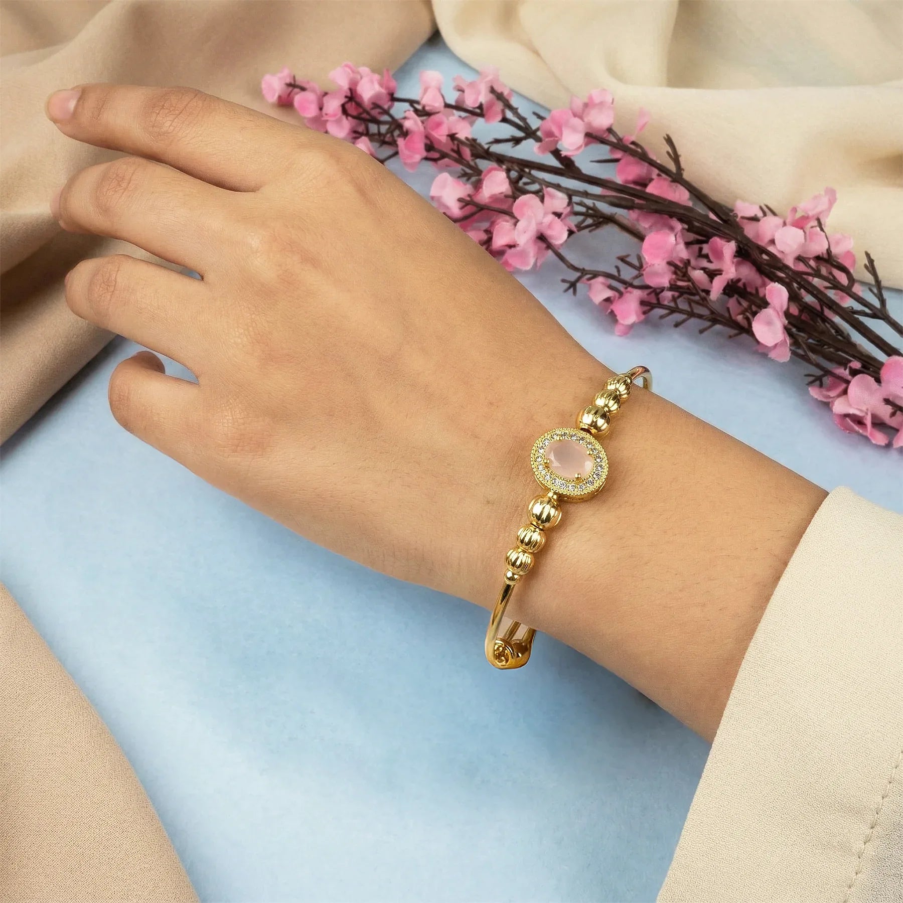 Gold bracelet with a pink gemstone on a wrist, with pink flowers in the background.