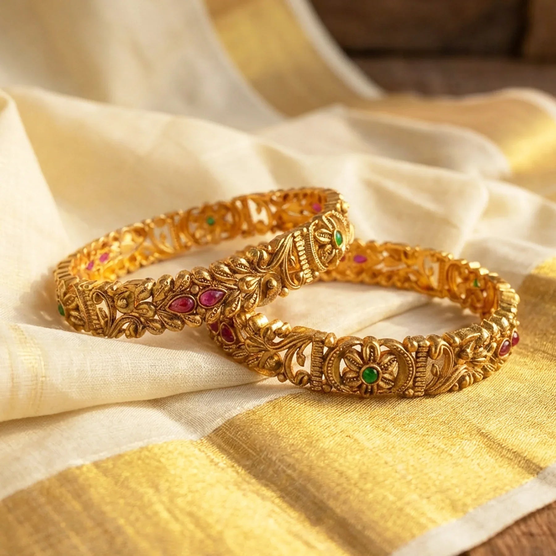 Two gold bangles with gemstones on a yellow and white fabric background