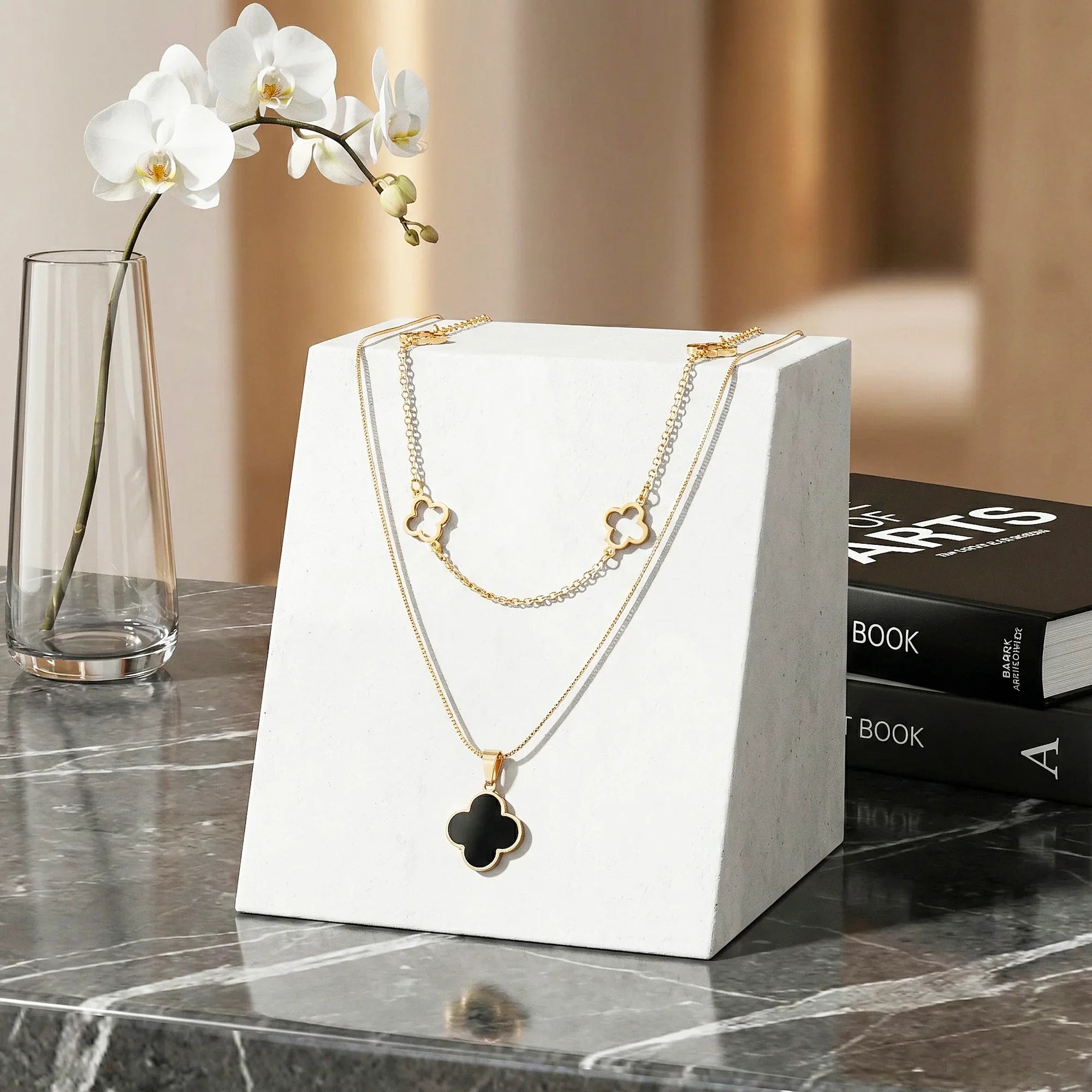 Gold necklace with a black clover pendant on a white stand, with a vase and books in the background.