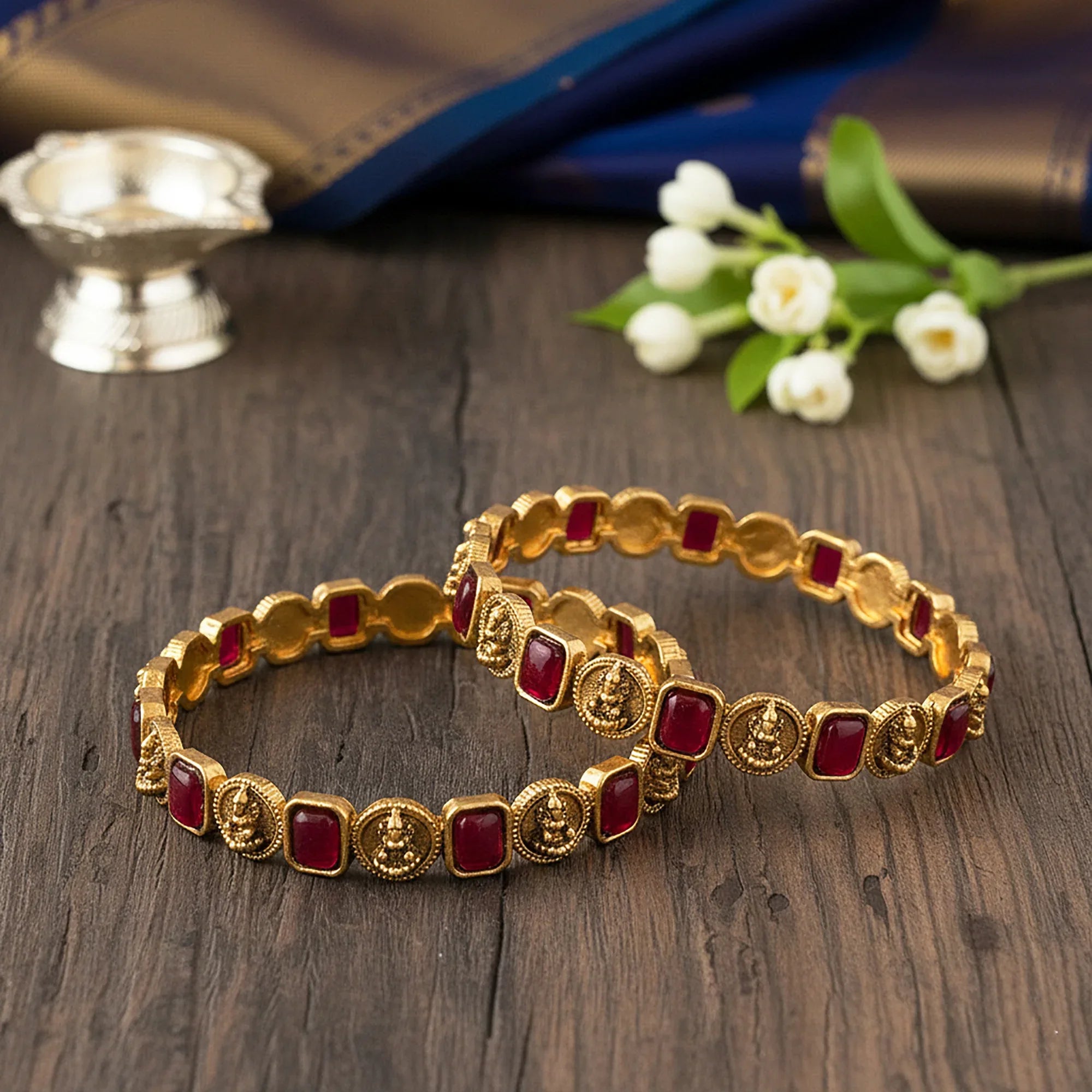 Gold bracelets with red stones on a wooden surface with flowers and a candle in the background