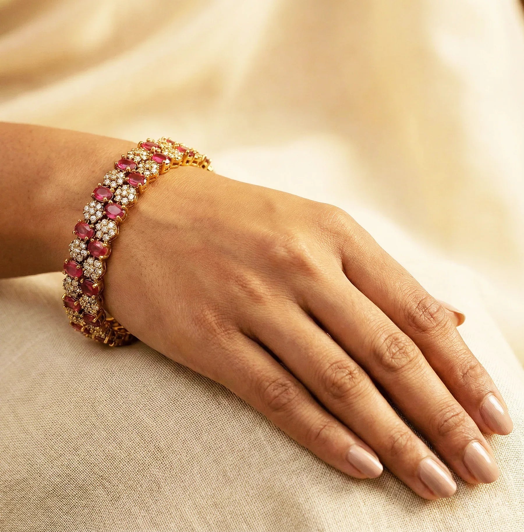 Hand wearing a gold bracelet with red gemstones on a beige fabric background