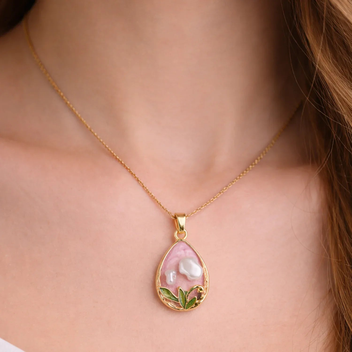 Gold necklace with a pink pendant featuring pearls and green leaves on a person's neck.