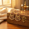 Gold earrings with pearl and red stone details on a wooden box with a warm glow.