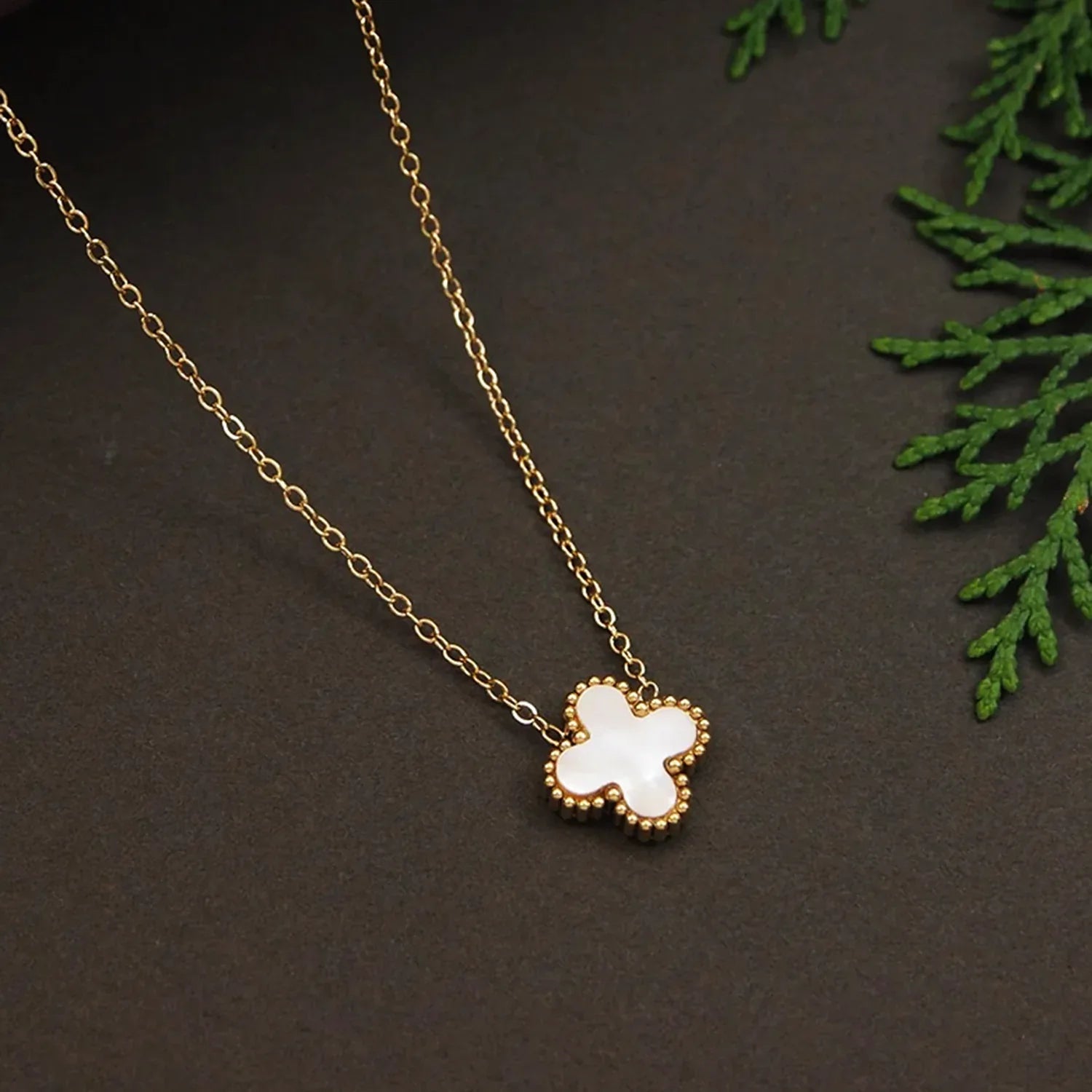 Gold necklace with a white flower pendant on a dark surface with green leaves.
