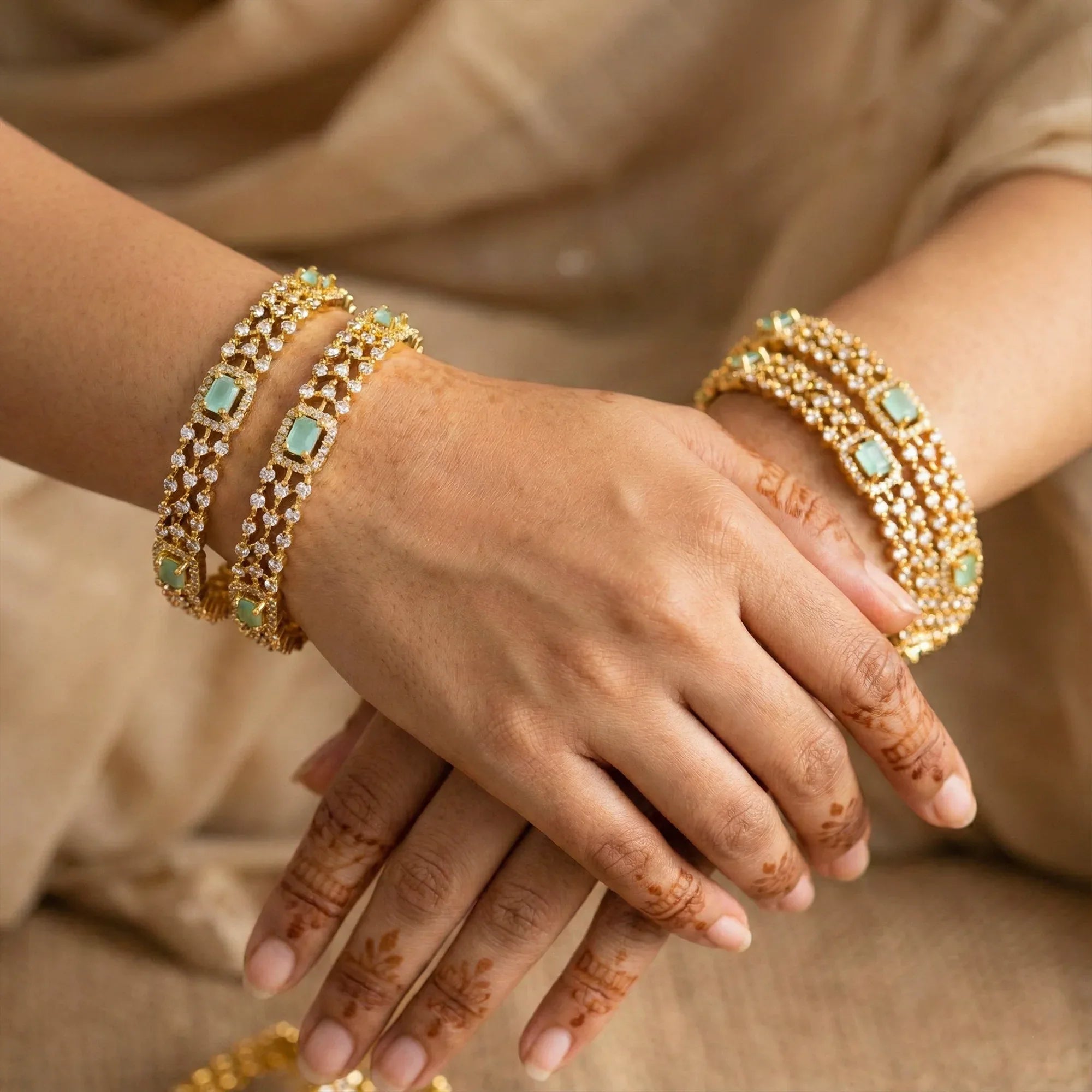 Close-up of hands wearing gold bracelets with green gemstones against a neutral background