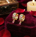 Gold hoop earrings with gemstones on a purple velvet stand with red rose petals and a candle in the background.