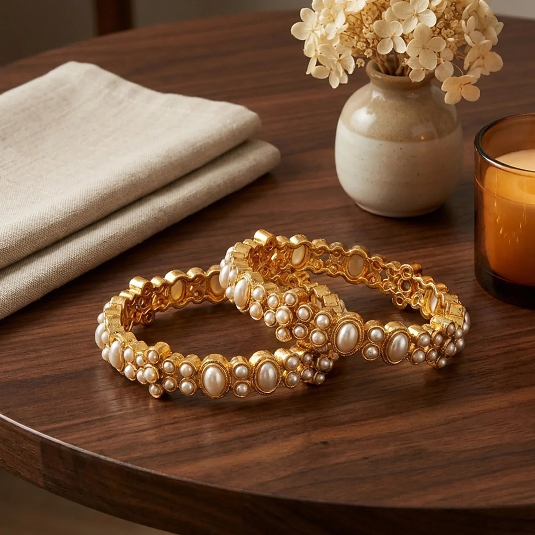 Two gold and pearl bracelets on a wooden table with a vase of flowers and a candle in the background.
