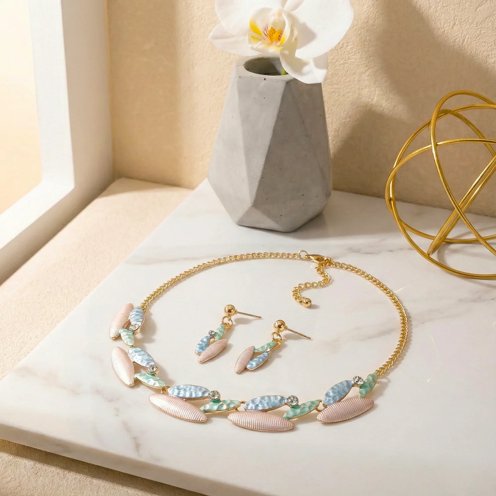 Jewelry set with necklace and earrings on a marble surface with a vase and decorative object in the background.