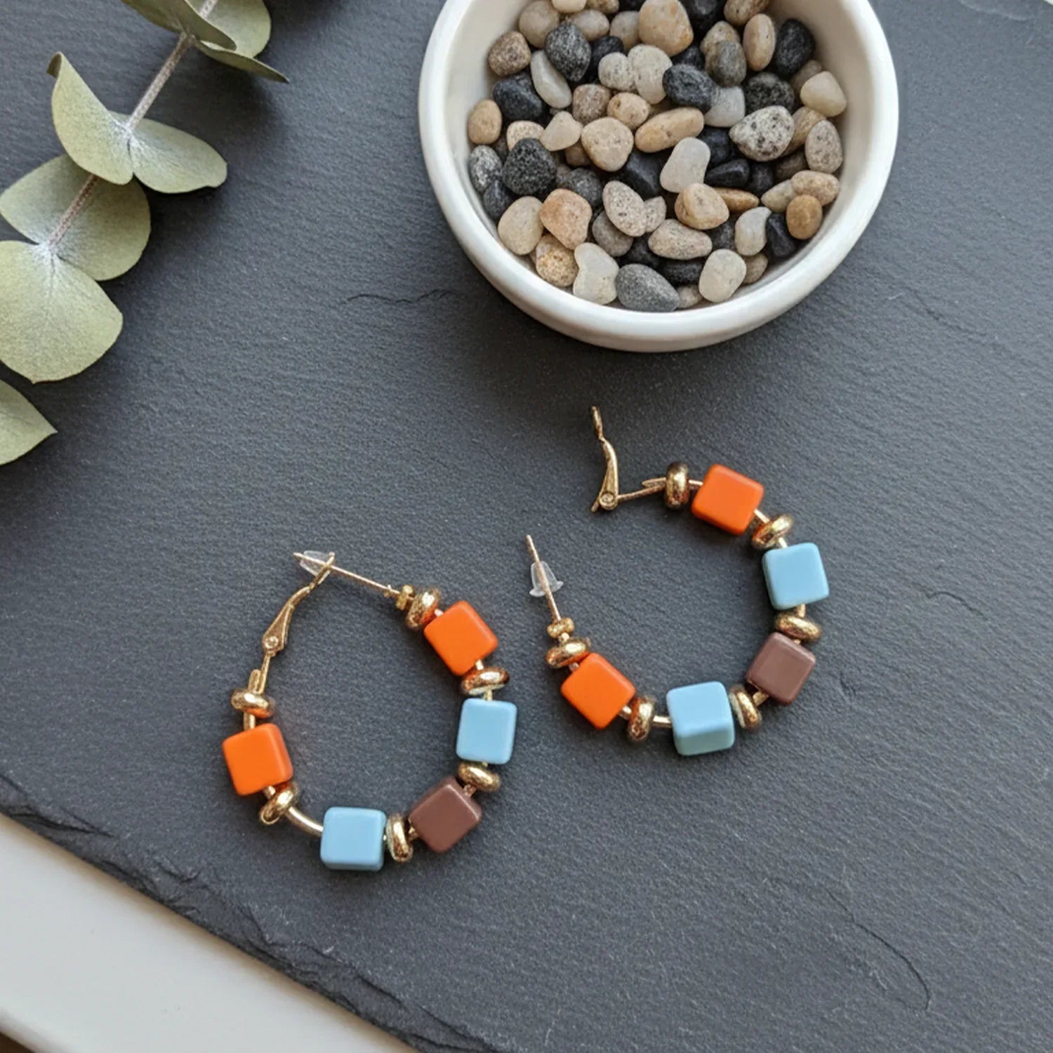 Colorful hoop earrings on a dark surface with pebbles and eucalyptus leaves.