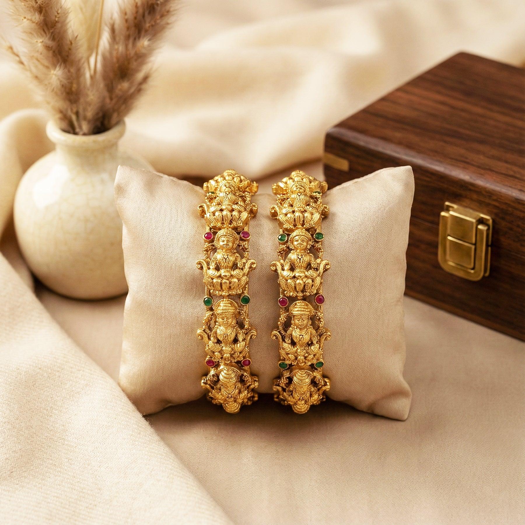 Pair of gold bangles with gemstones on a beige pillow with a wooden box in the background.
