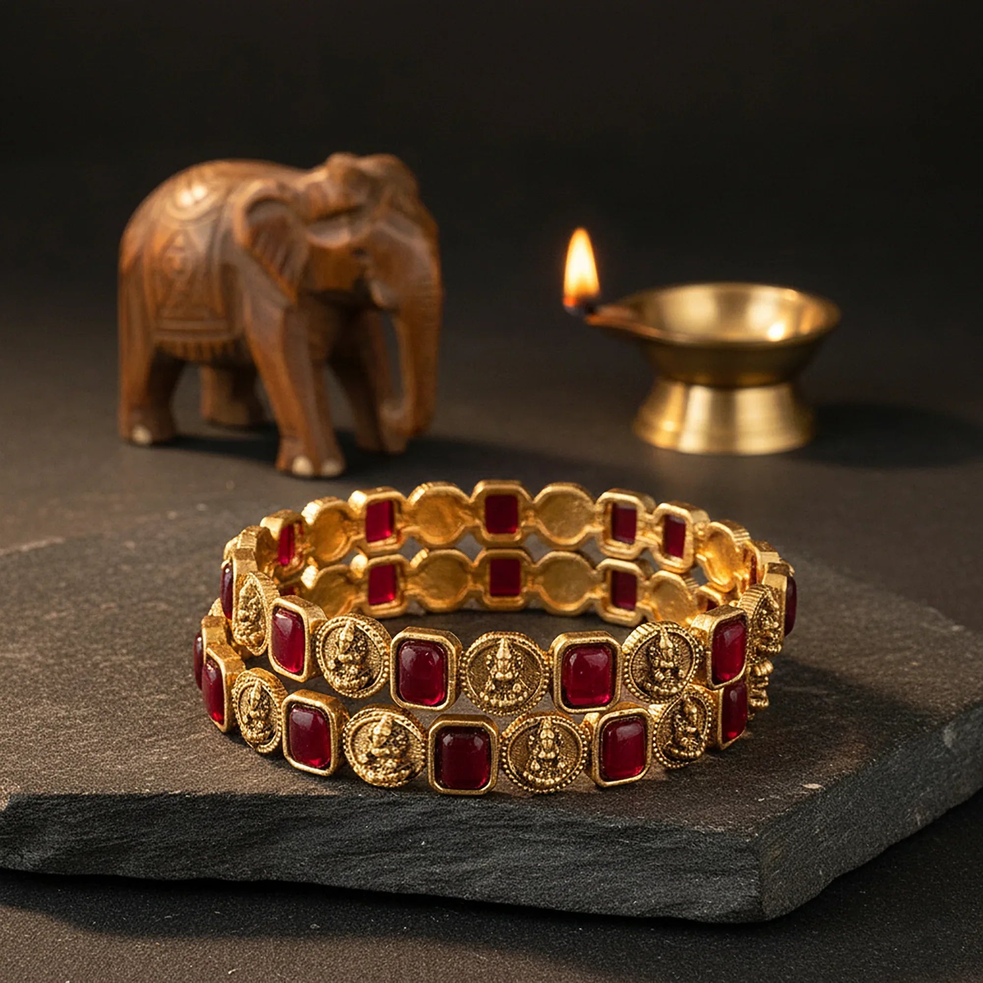 Gold and red bracelet on a dark surface with an elephant and lit candle in the background