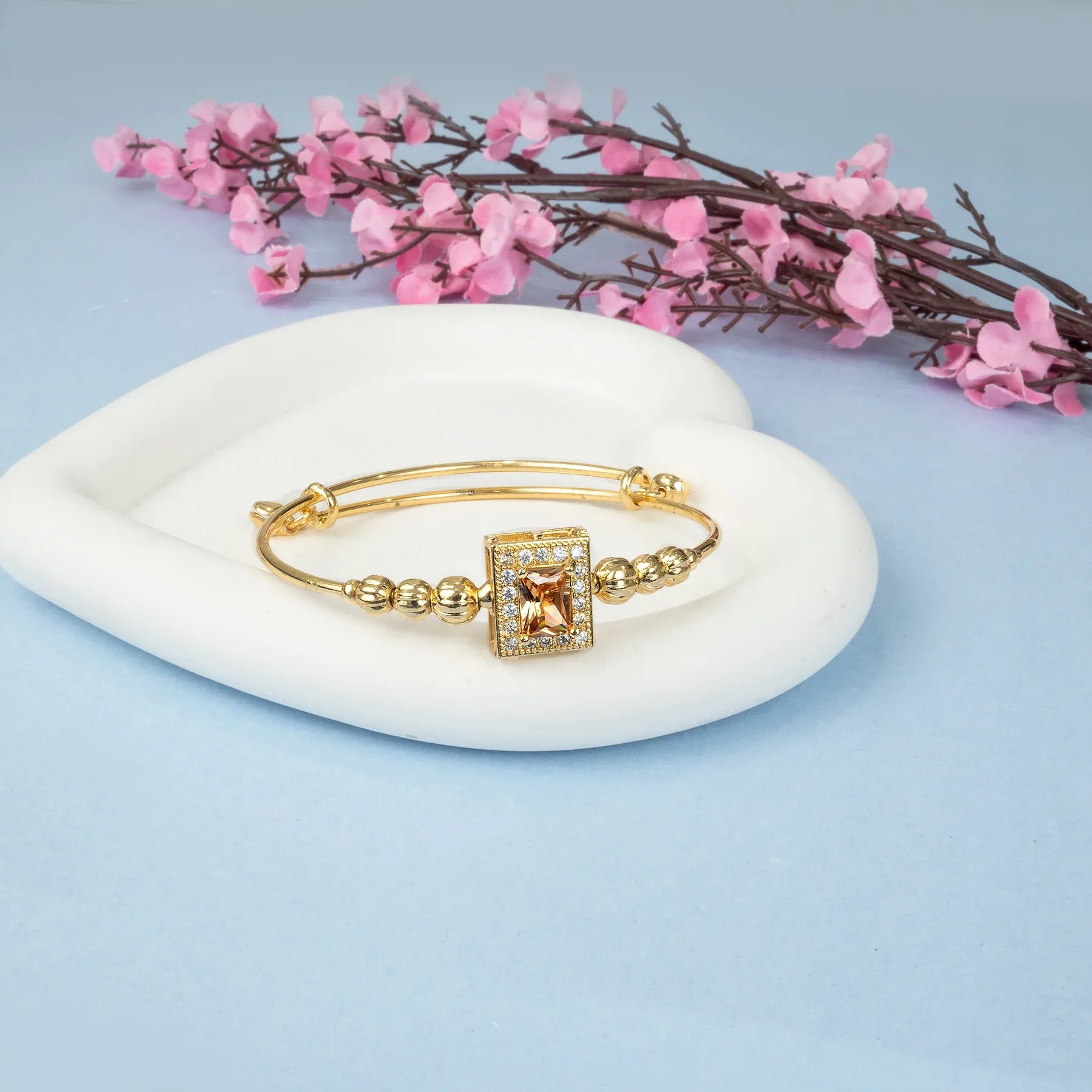 Gold bracelet with a square pendant on a white dish with pink flowers in the background