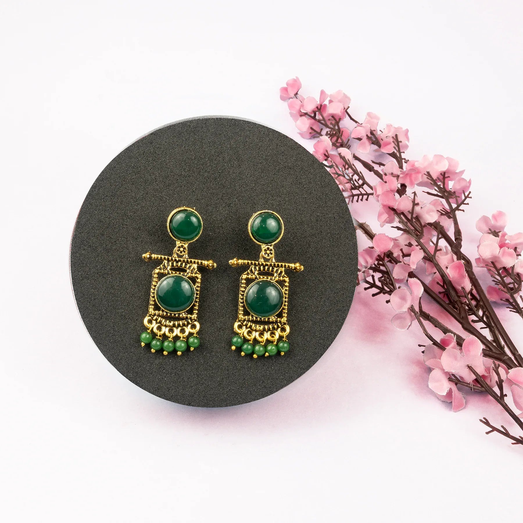 Pair of green and gold earrings on a black stand with pink flowers in the background