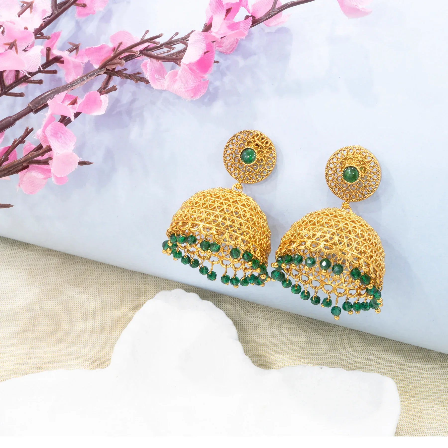 Gold earrings with green stones on a white surface with pink flowers in the background