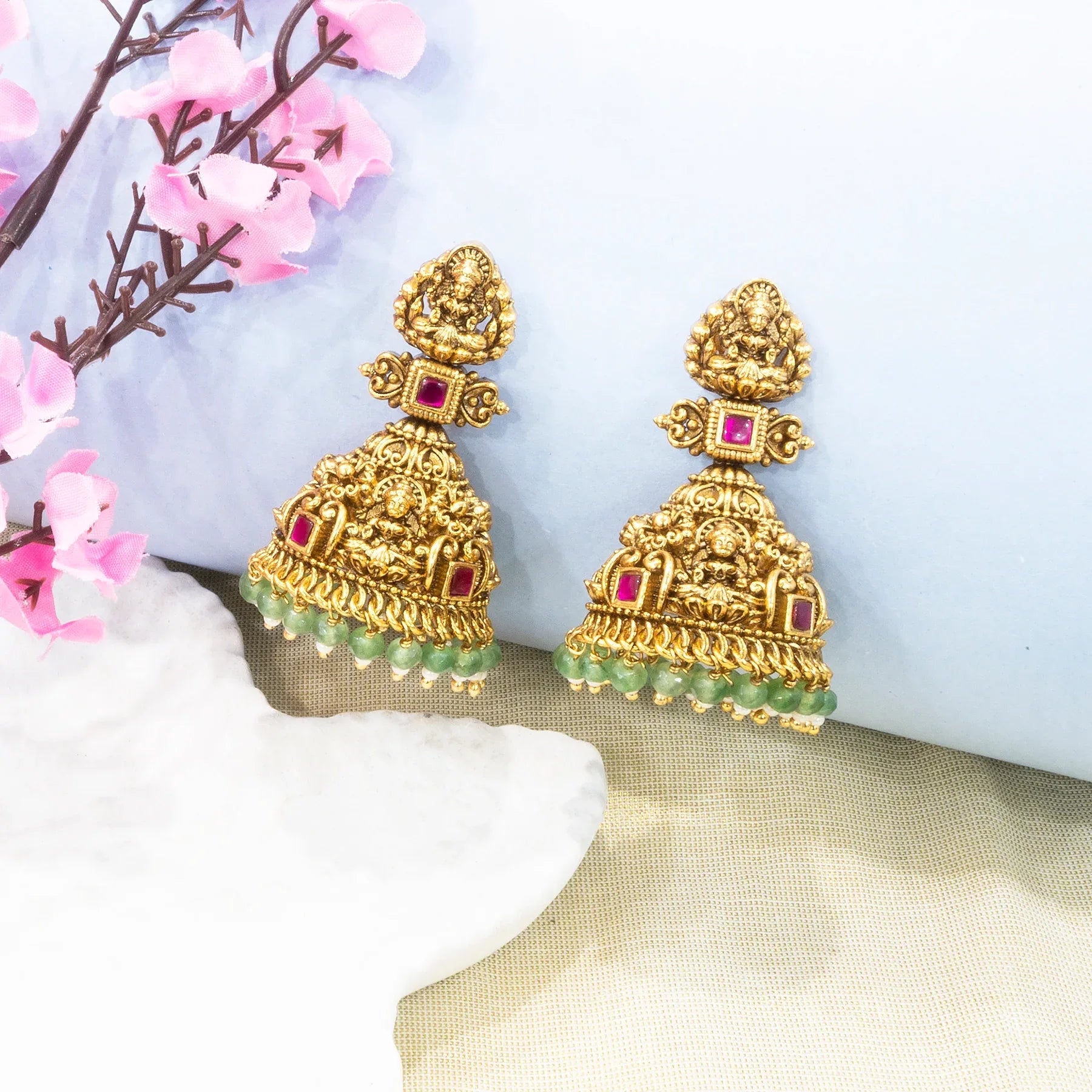 Gold earrings with red and green stones on a soft white background with pink flowers.