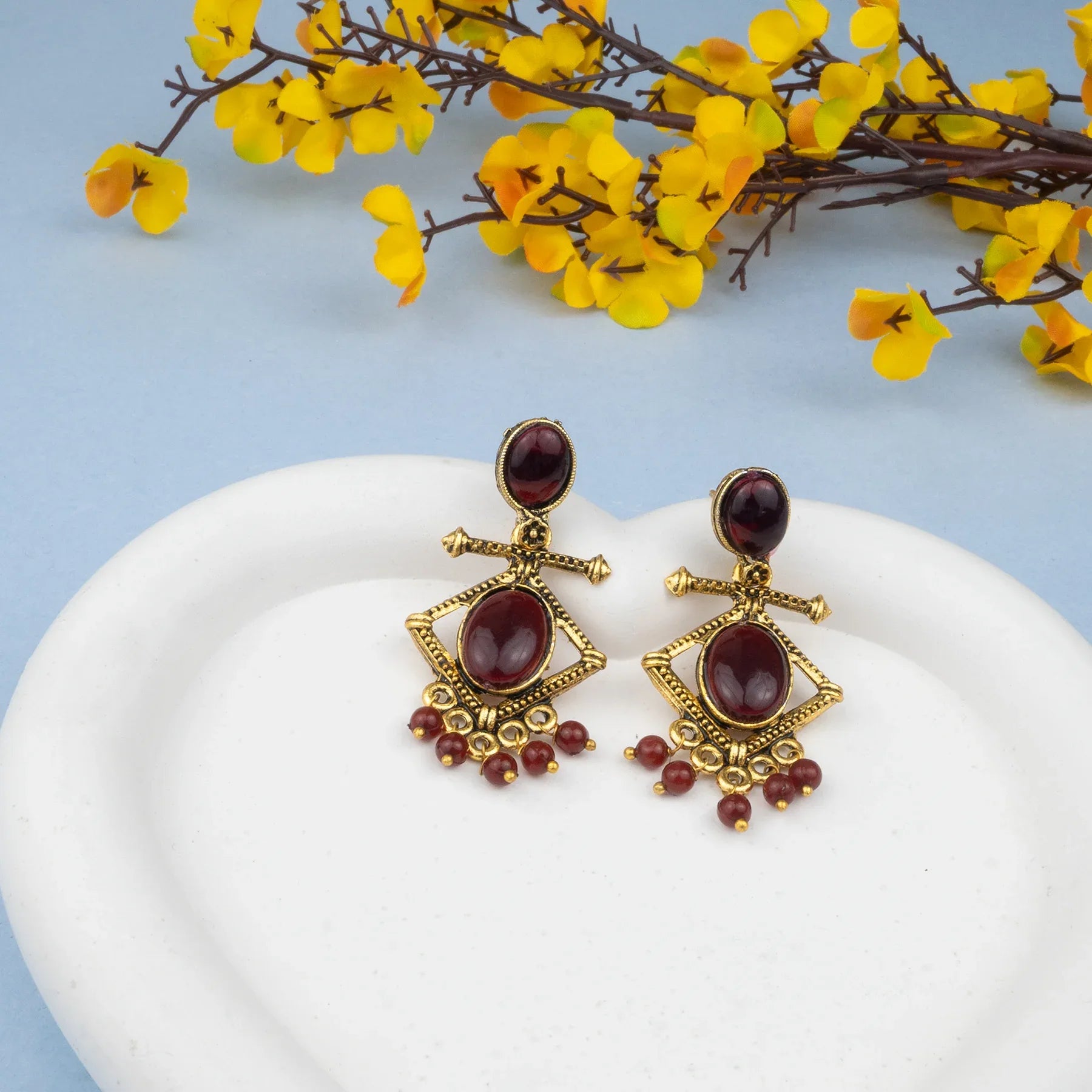Pair of gold and red earrings on a white heart-shaped object with yellow flowers in the background.