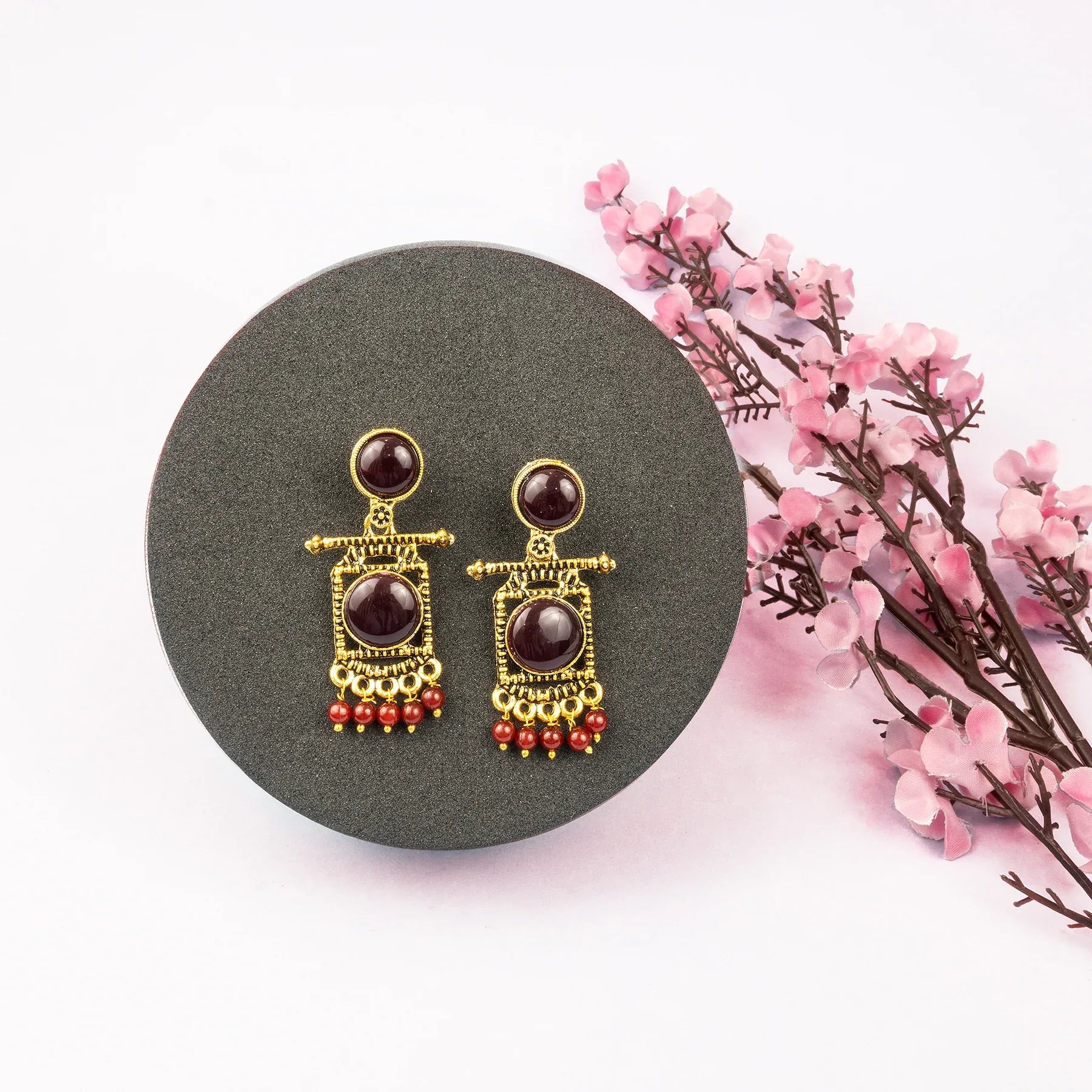 Pair of gold and red earrings on a gray stand with pink flowers in the background