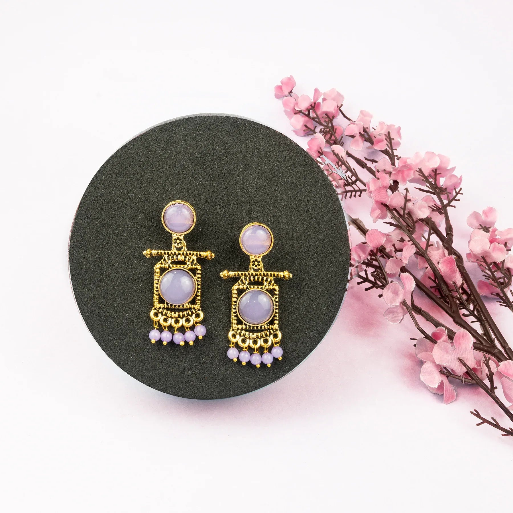 Gold earrings with pink stones on a black circular stand with pink flowers in the background