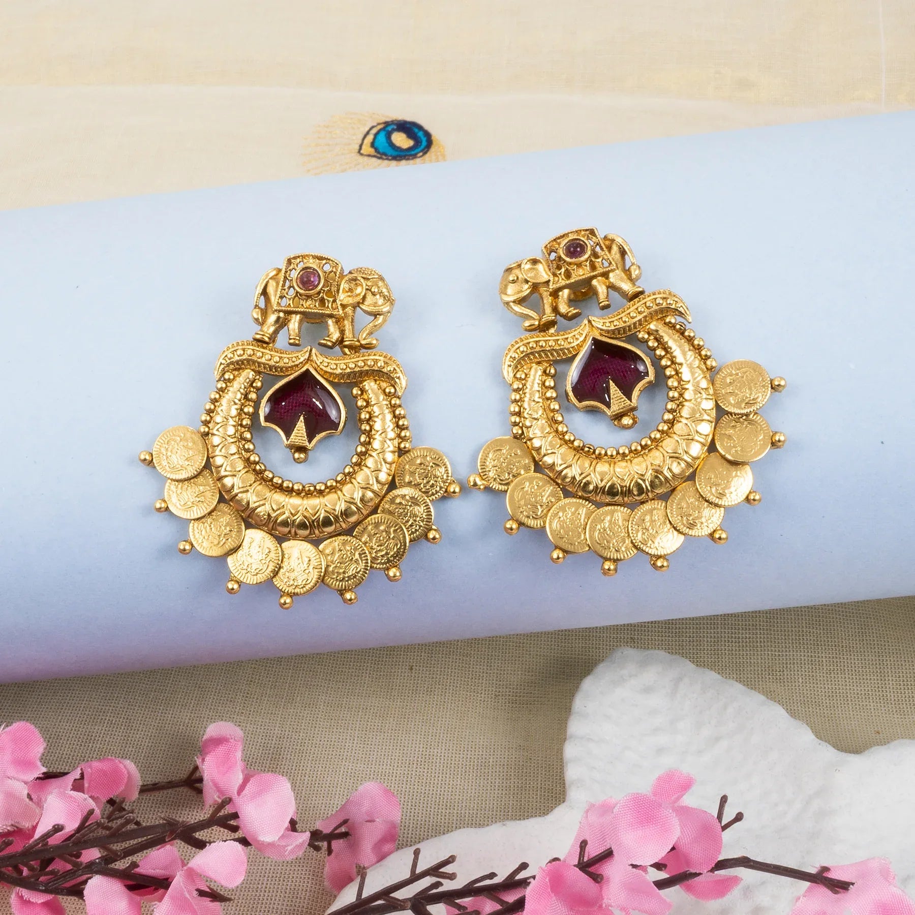 Gold earrings with red gemstones on a light blue surface with pink flowers in the background