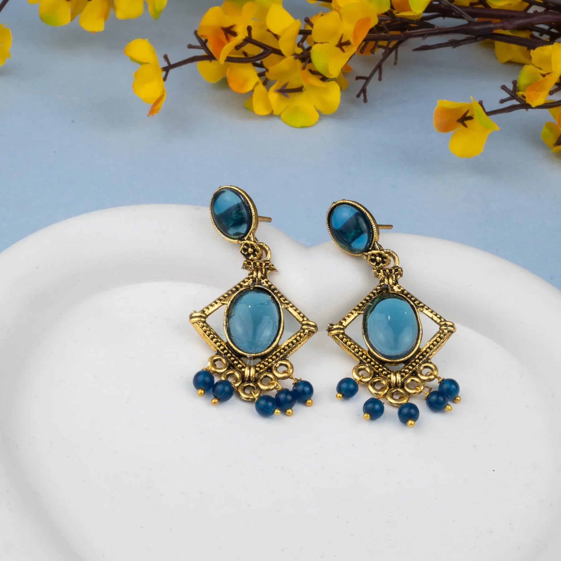 Gold earrings with blue stones on a white surface with yellow flowers in the background