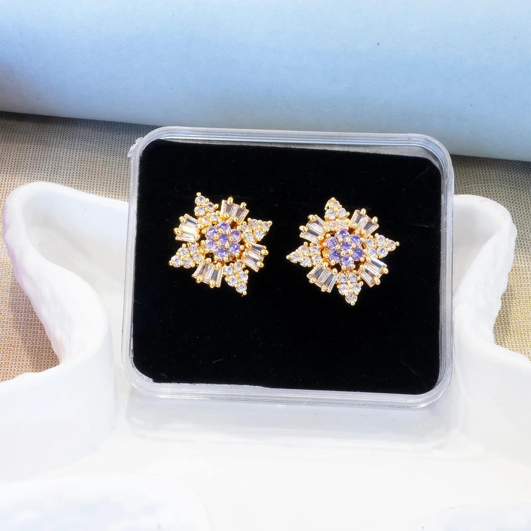 Pair of gold and purple star-shaped earrings in a clear box on a beige surface.
