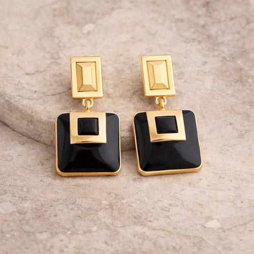 Gold and black square-shaped earrings on a beige stone background