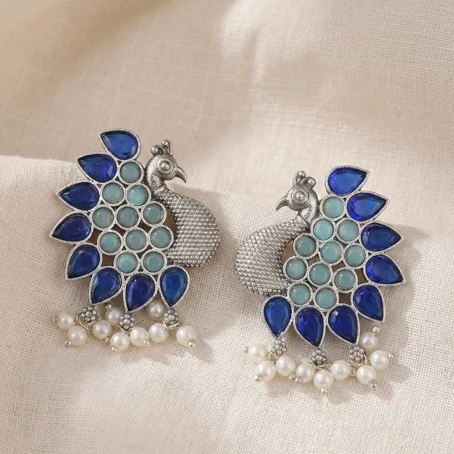 Pair of peacock-shaped earrings with blue and green stones on a beige fabric background