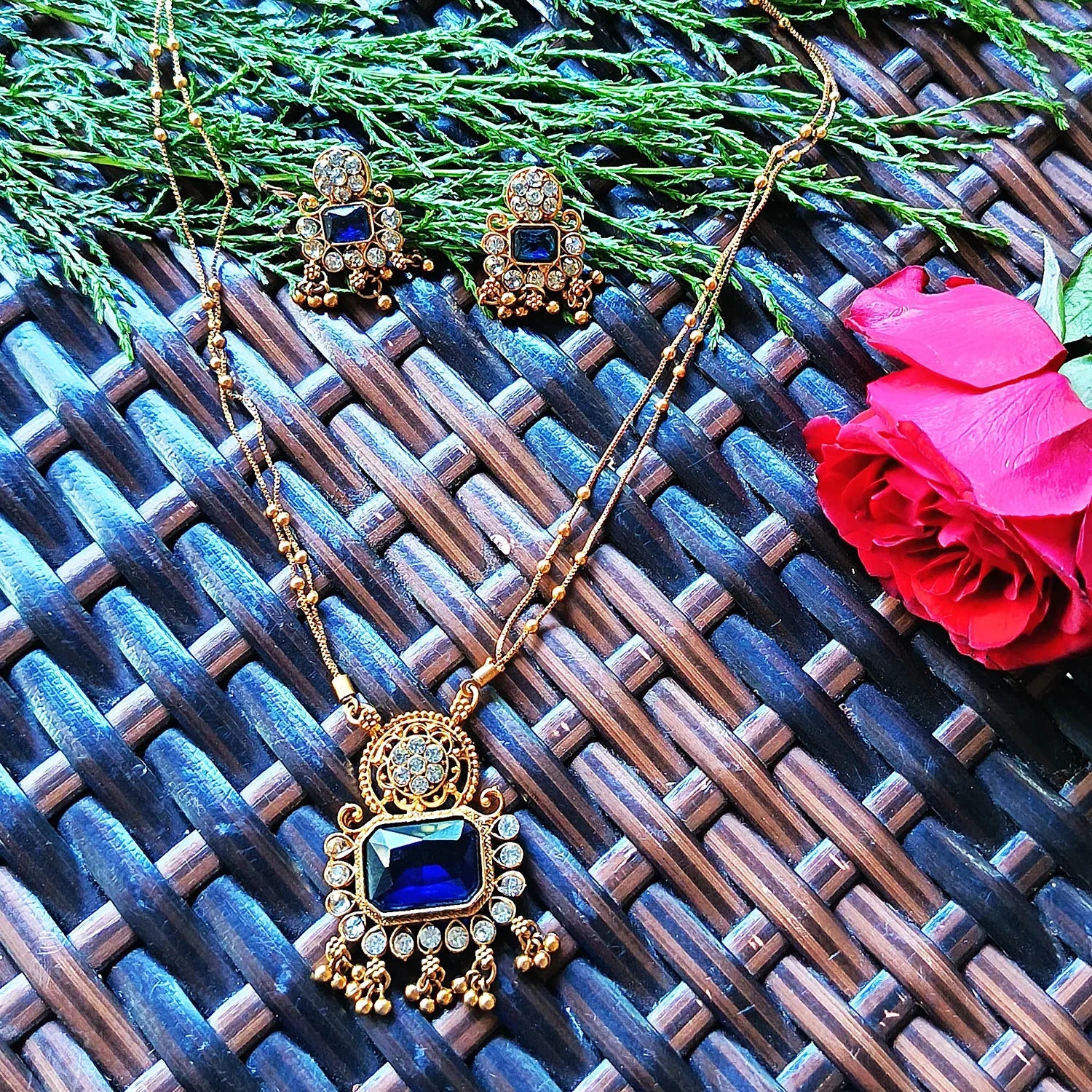 Gold necklace with blue gemstone and matching earrings on a textured surface with a pink rose.