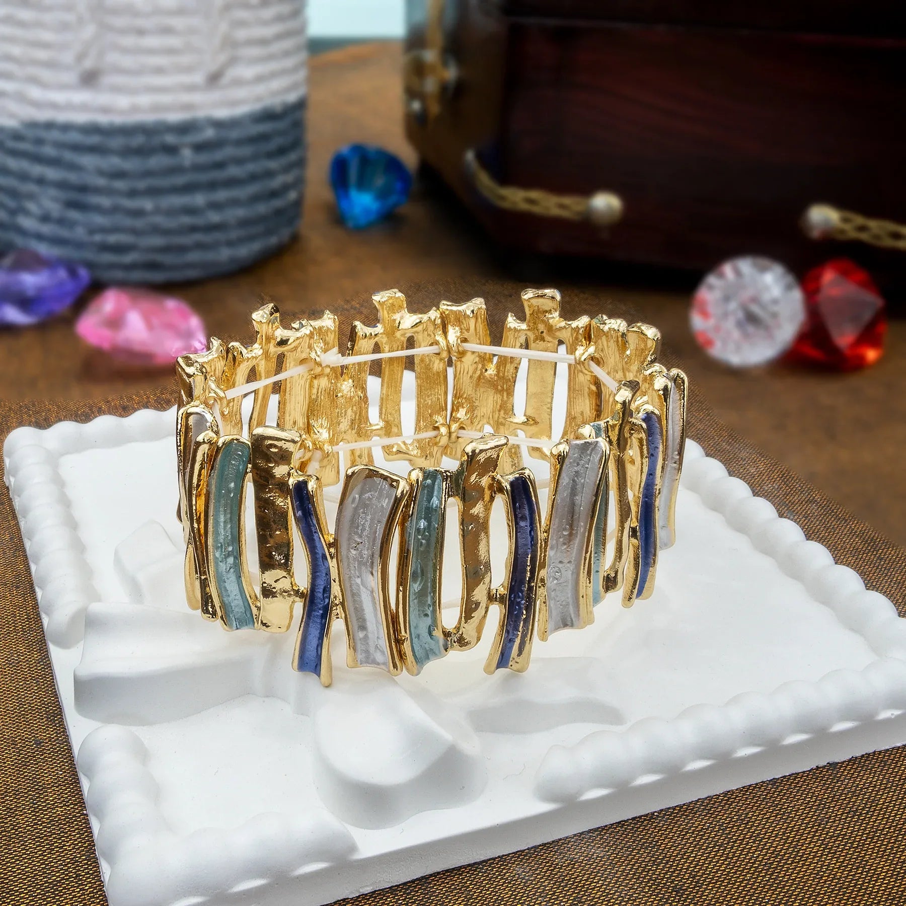 Gold bracelet with colorful stones on a white stand against a textured surface.