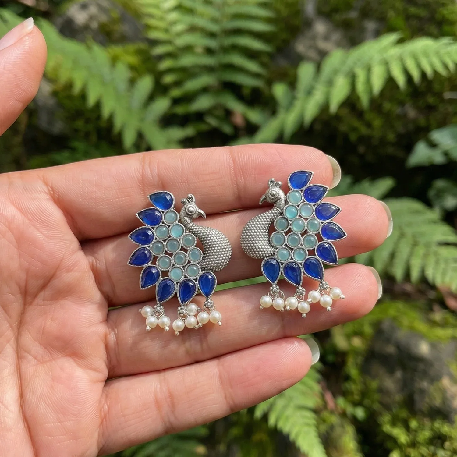 Pair of peacock-shaped earrings with blue and green stones held between fingers against a natural background.
