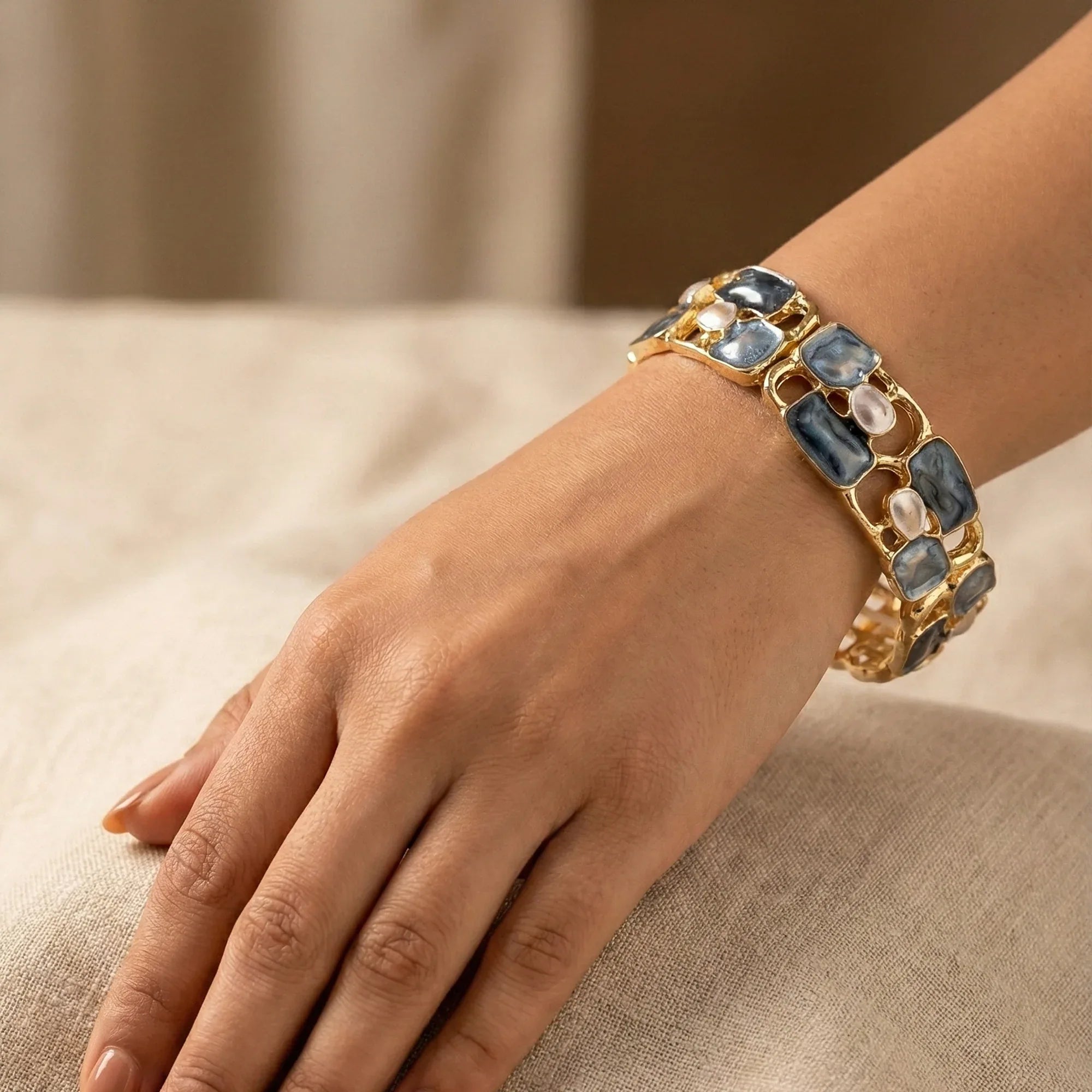 Hand wearing a gold bracelet with blue stones on a beige fabric background