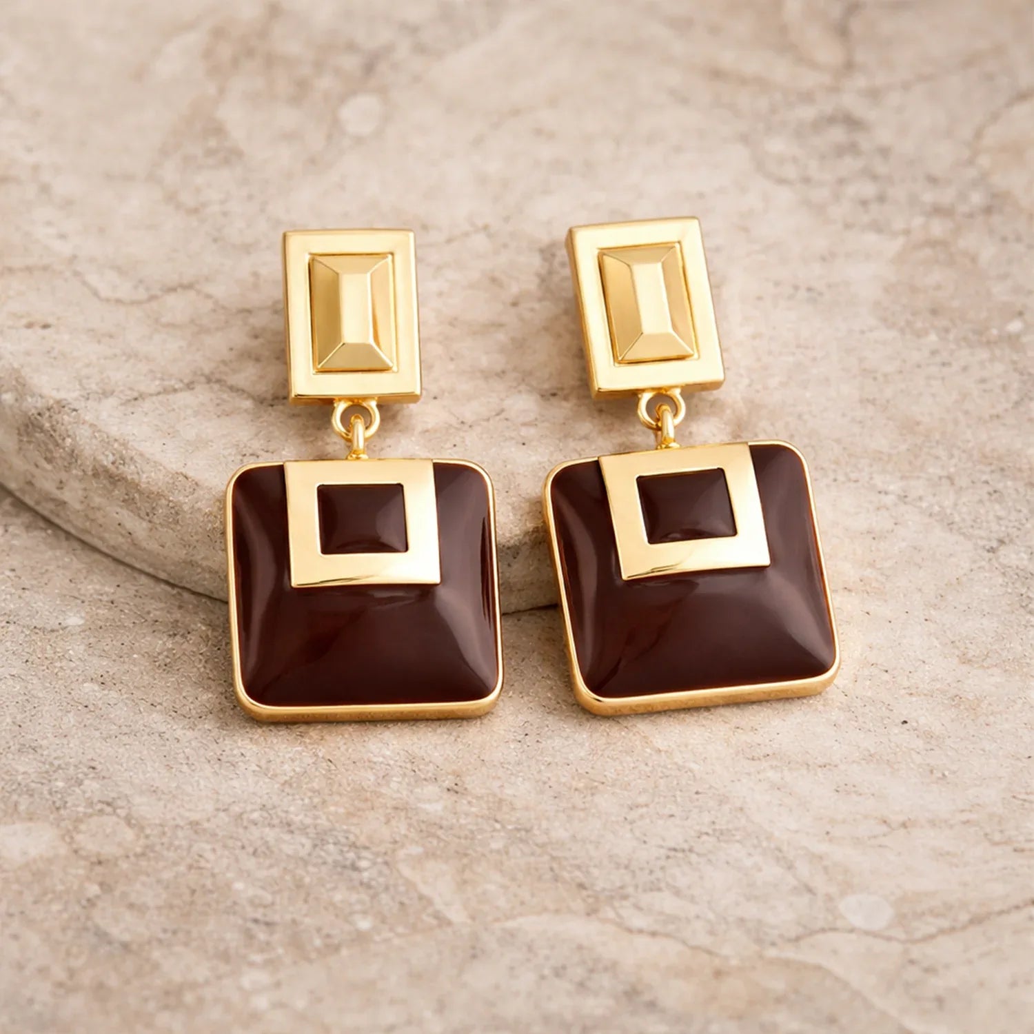Gold and brown square earrings on a beige stone surface
