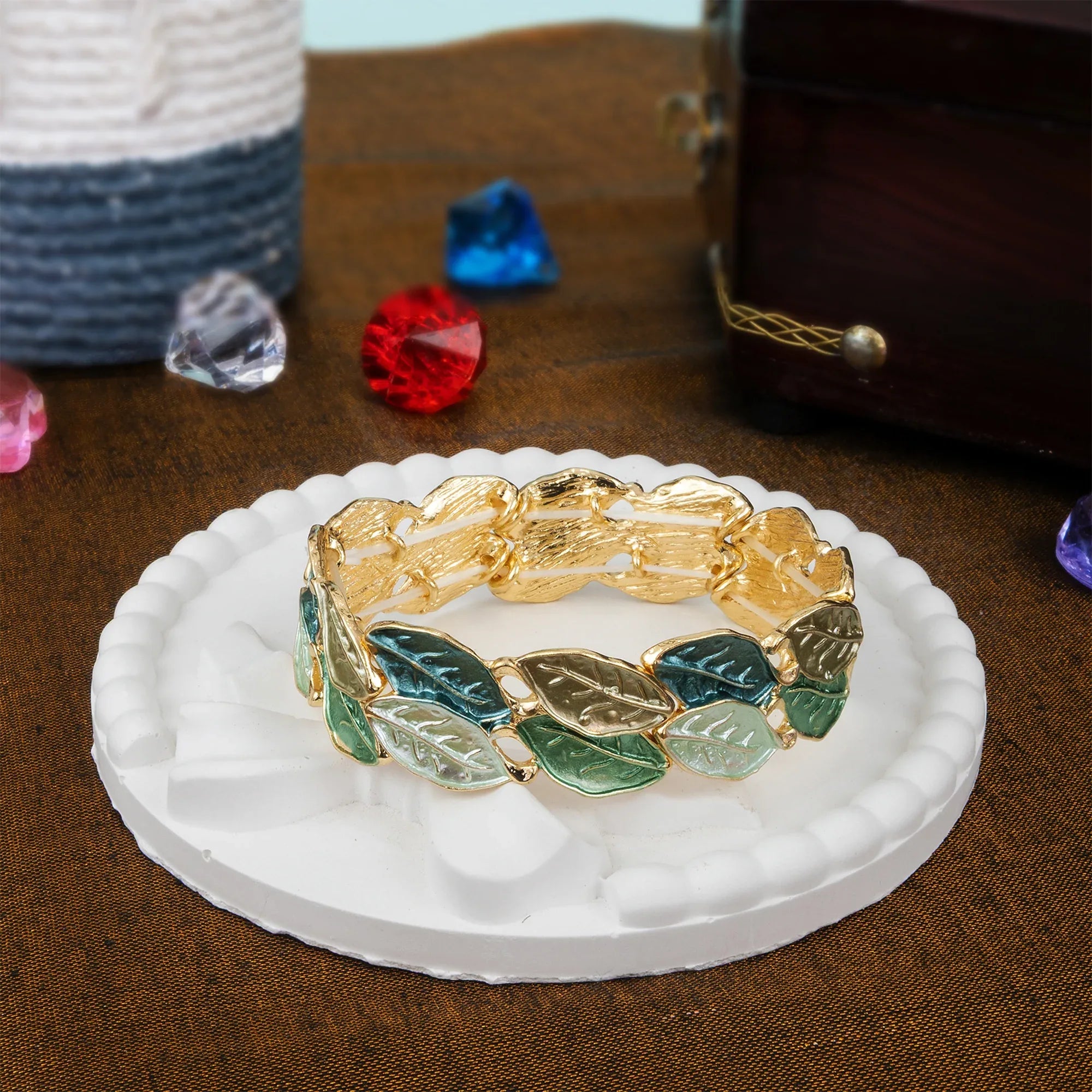 Gold bracelet with leaf designs on a white stand, surrounded by colorful gemstones on a brown surface.