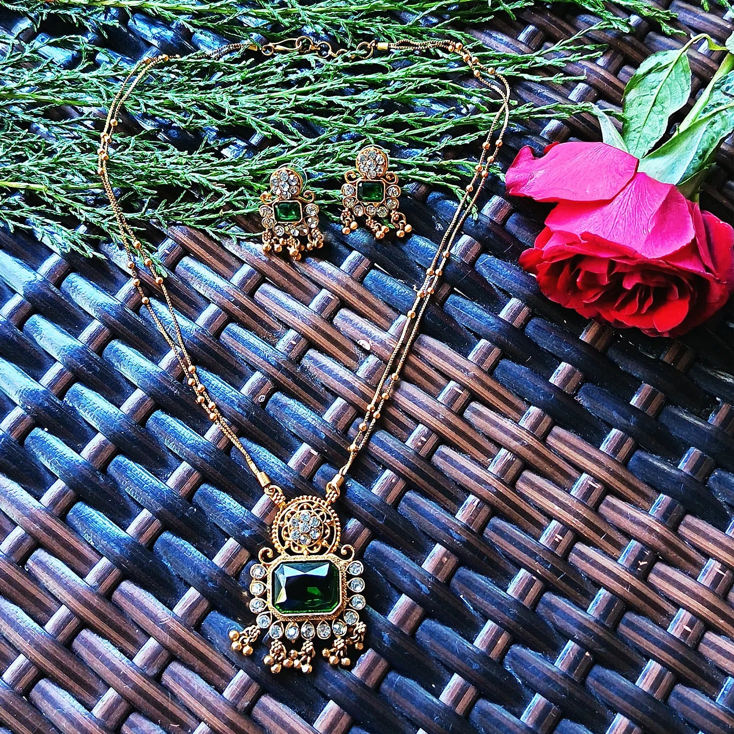 Gold necklace with green pendant and matching earrings on a textured surface with a red rose.