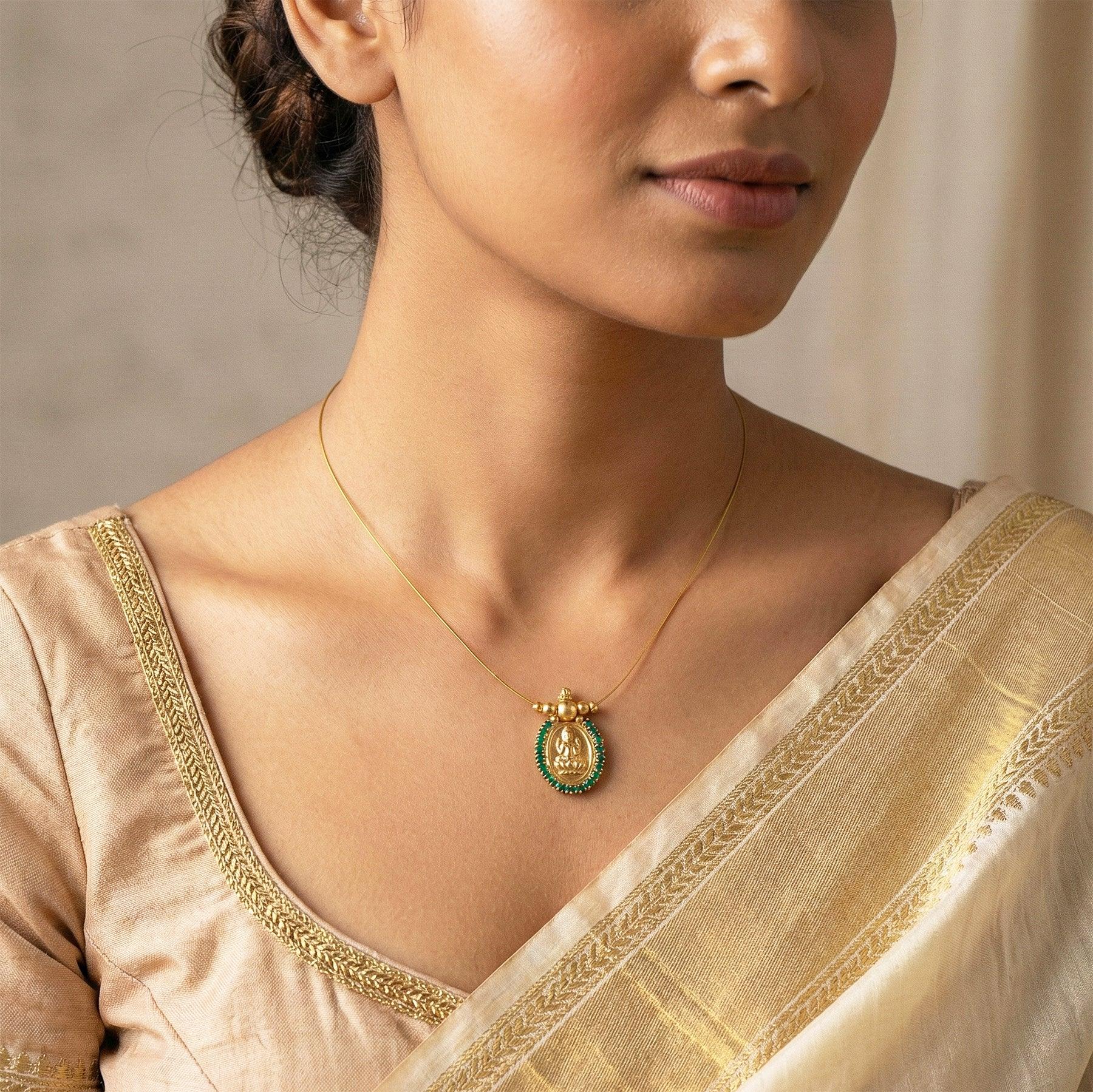 Woman wearing a gold necklace with a green pendant, dressed in a beige saree.