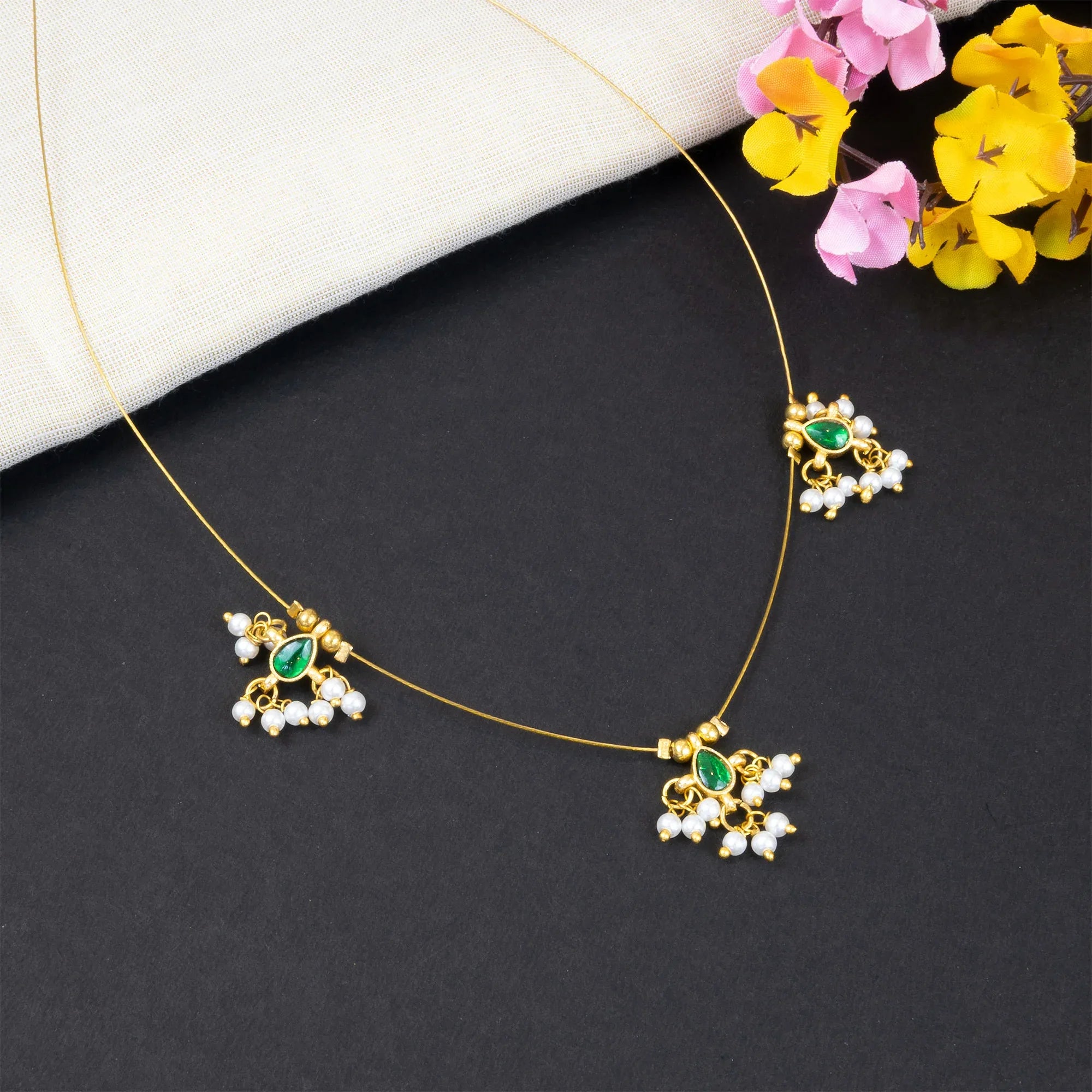 Gold necklace with green and white stones on a dark surface with flowers in the background