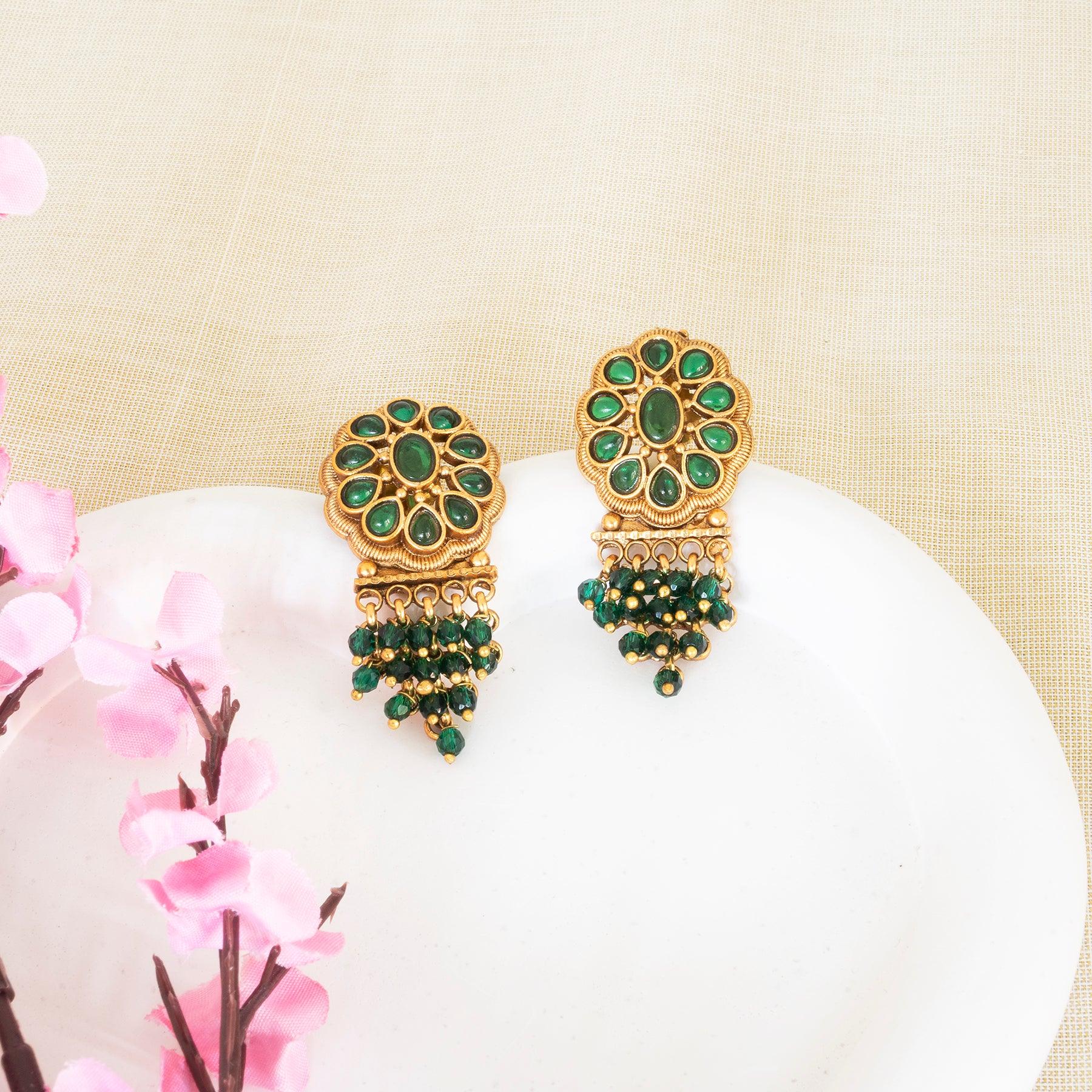 Gold and green earrings on a white surface with pink flowers in the background