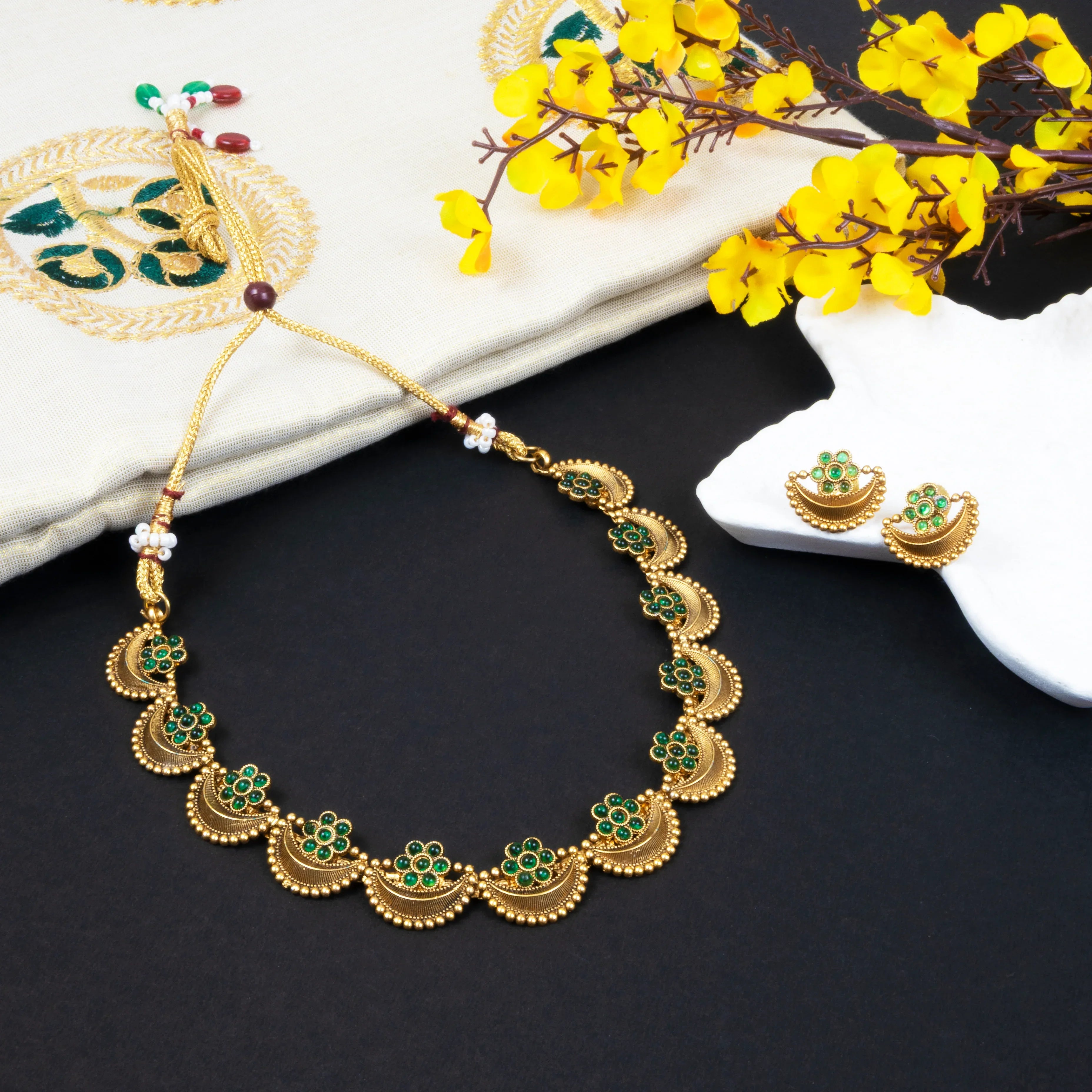 Gold necklace with green gemstones on a dark surface with yellow flowers.
