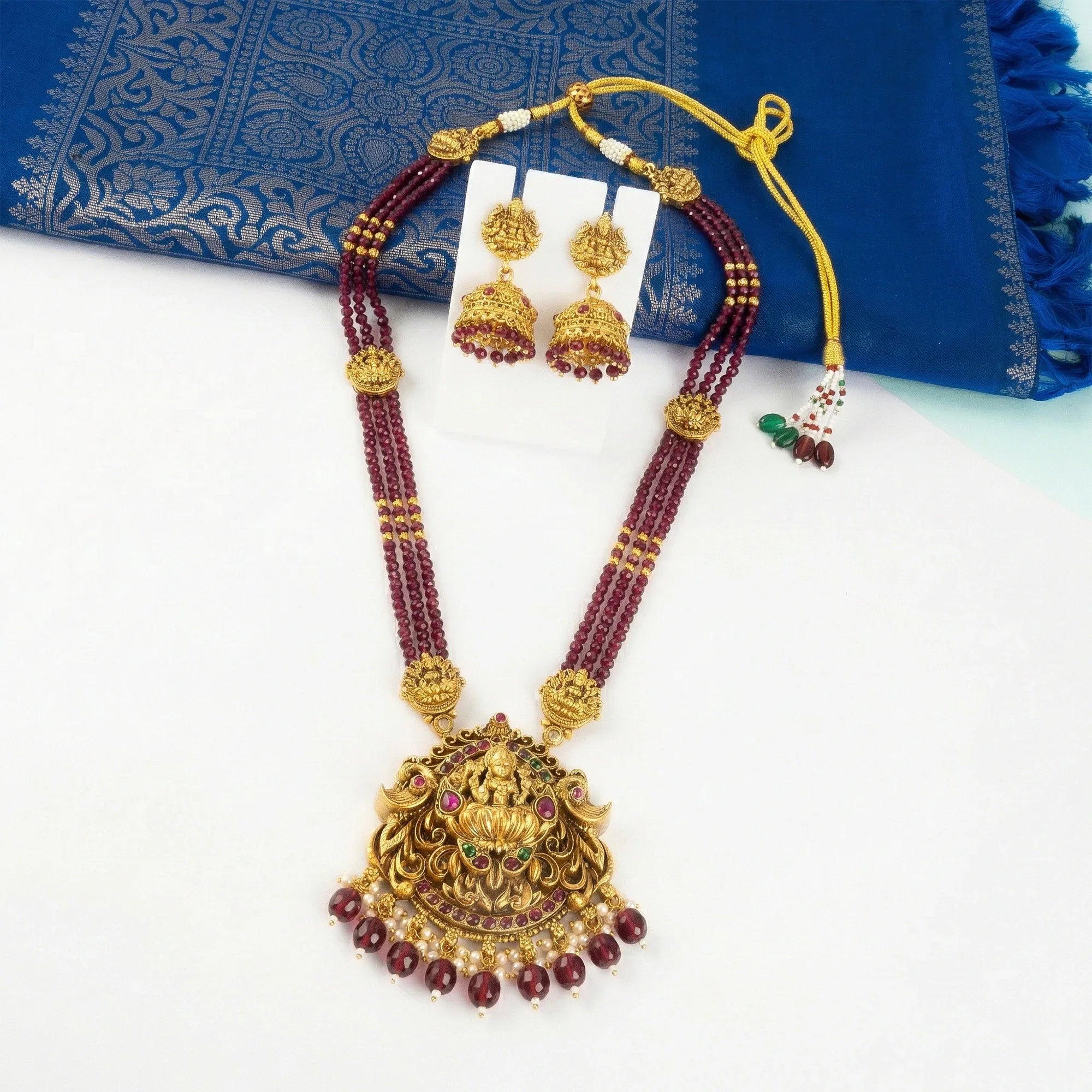 Gold necklace with maroon beads and matching earrings on a blue fabric background