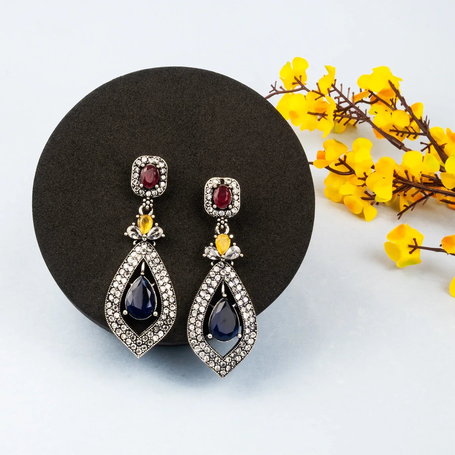 Pair of ornate earrings with gemstones on a black stand with yellow flowers in the background