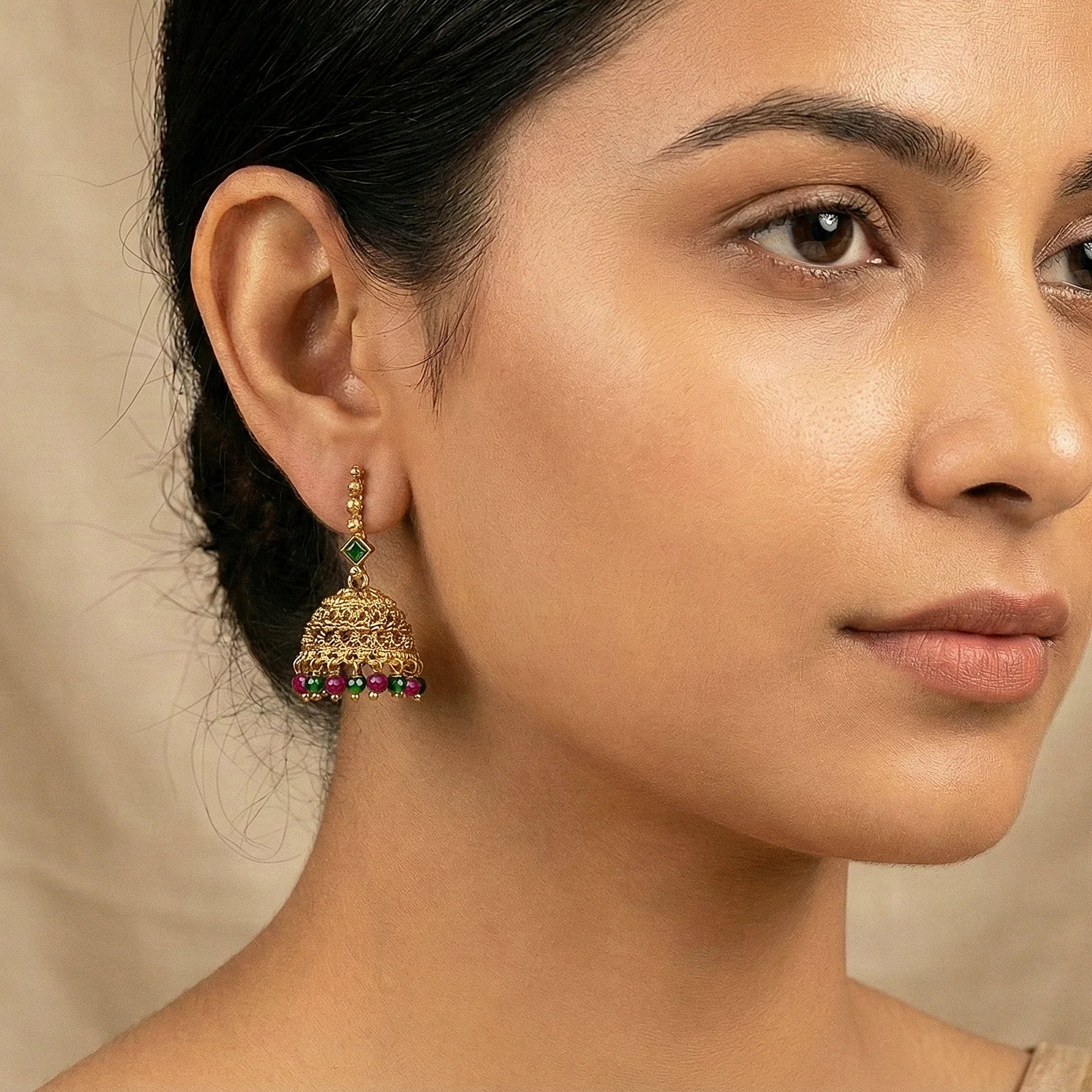 Close-up of a woman wearing gold earrings with red and green beads against a beige background