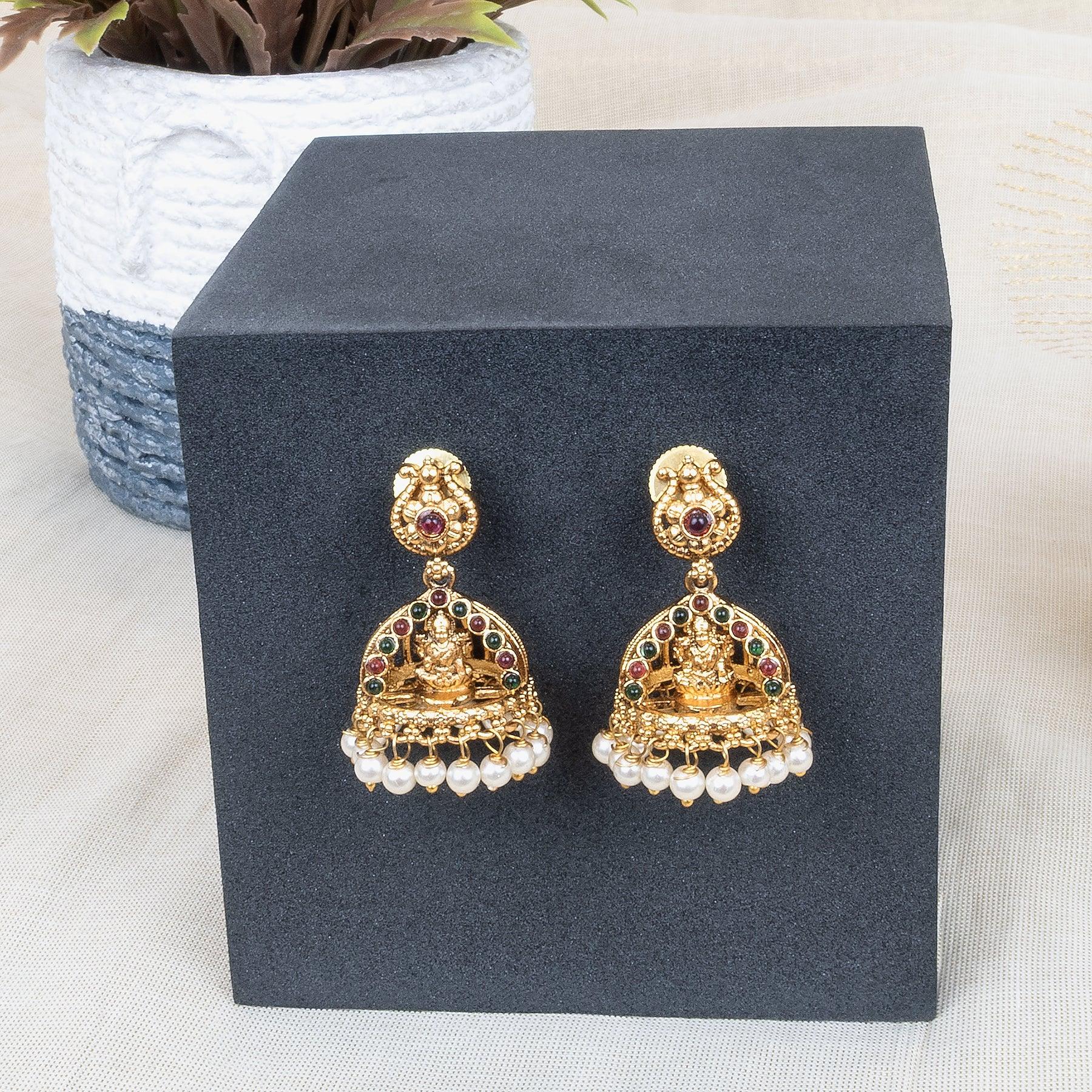 Gold earrings with red and green gemstones on a black stand with a plant in the background.