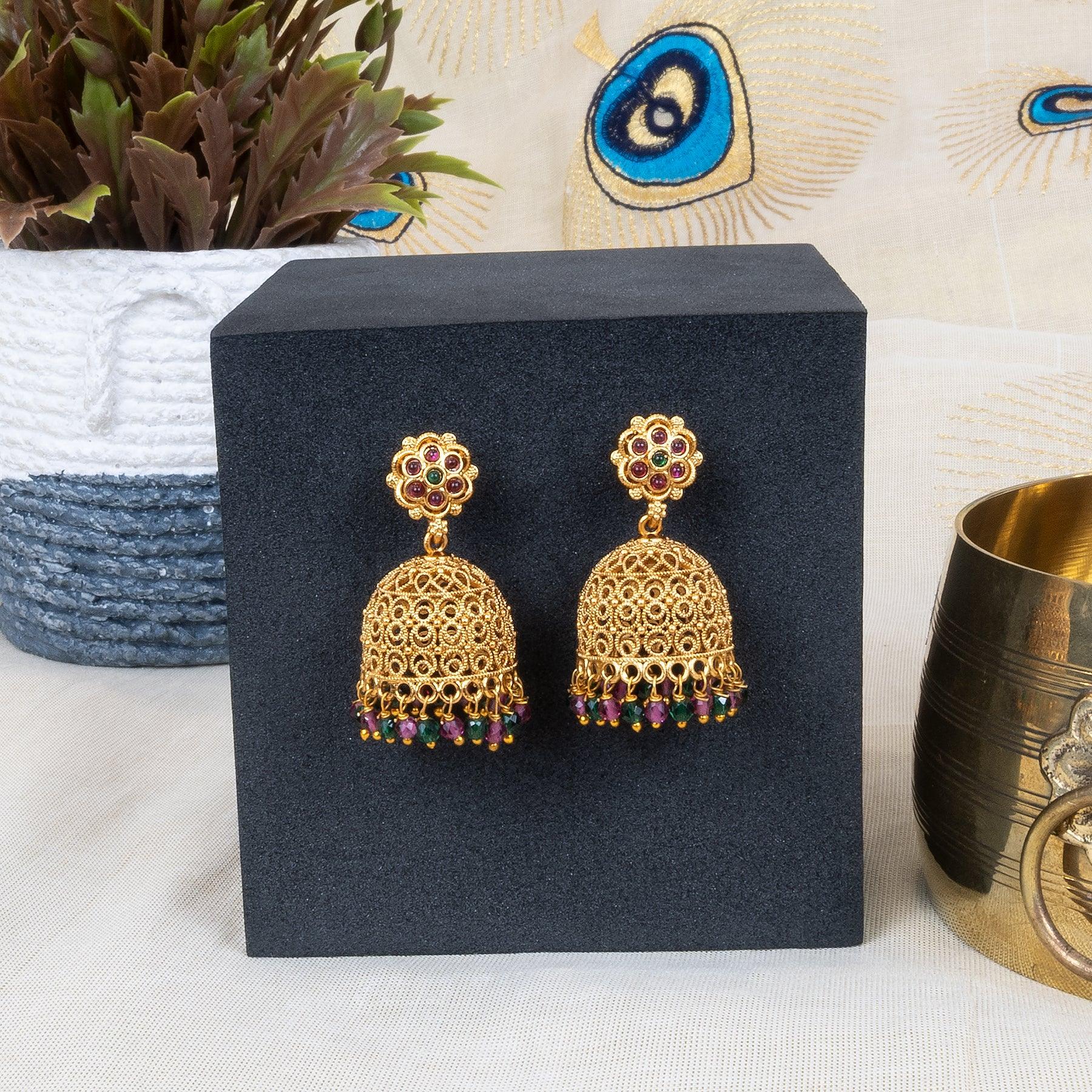 Gold earrings with colorful stones on a black stand, with a plant and decorative item in the background.