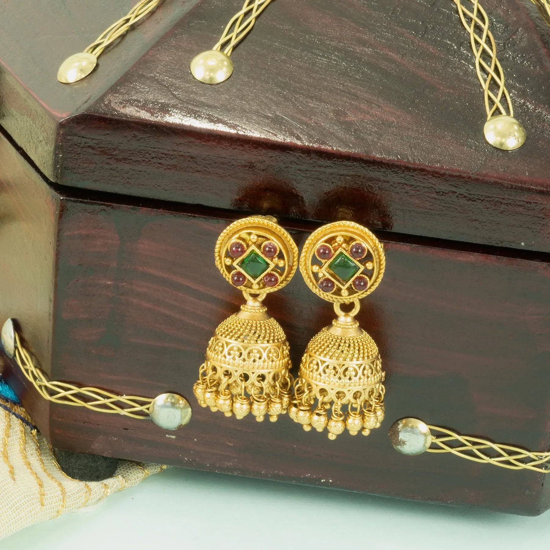 Gold earrings with green and red stones on a wooden jewelry box.