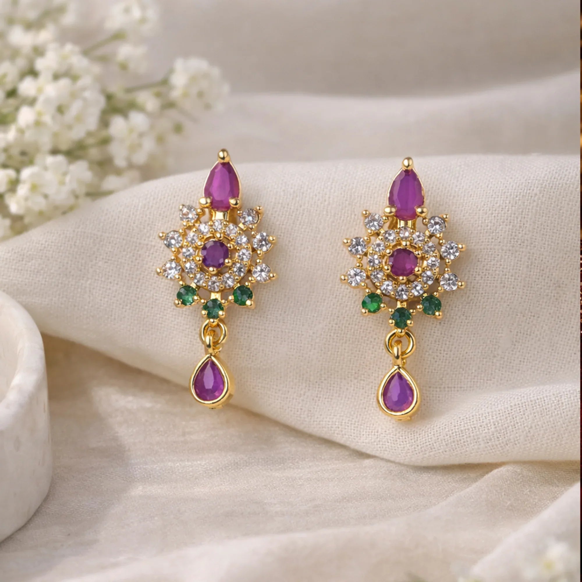 Gold earrings with purple and green gemstones on a beige surface with white flowers in the background