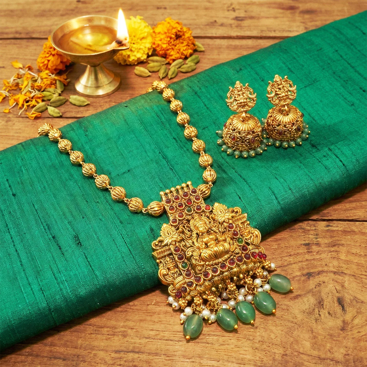 Gold necklace and earrings on a green fabric with flowers and oil lamp in the background