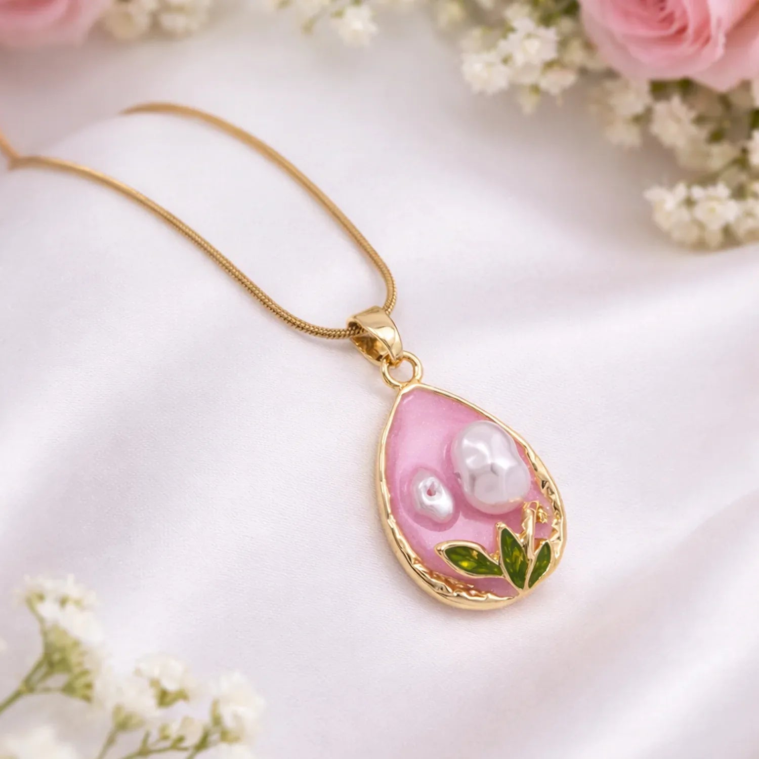 Gold necklace with a pink teardrop pendant featuring pearls and leaves on a white background with flowers.