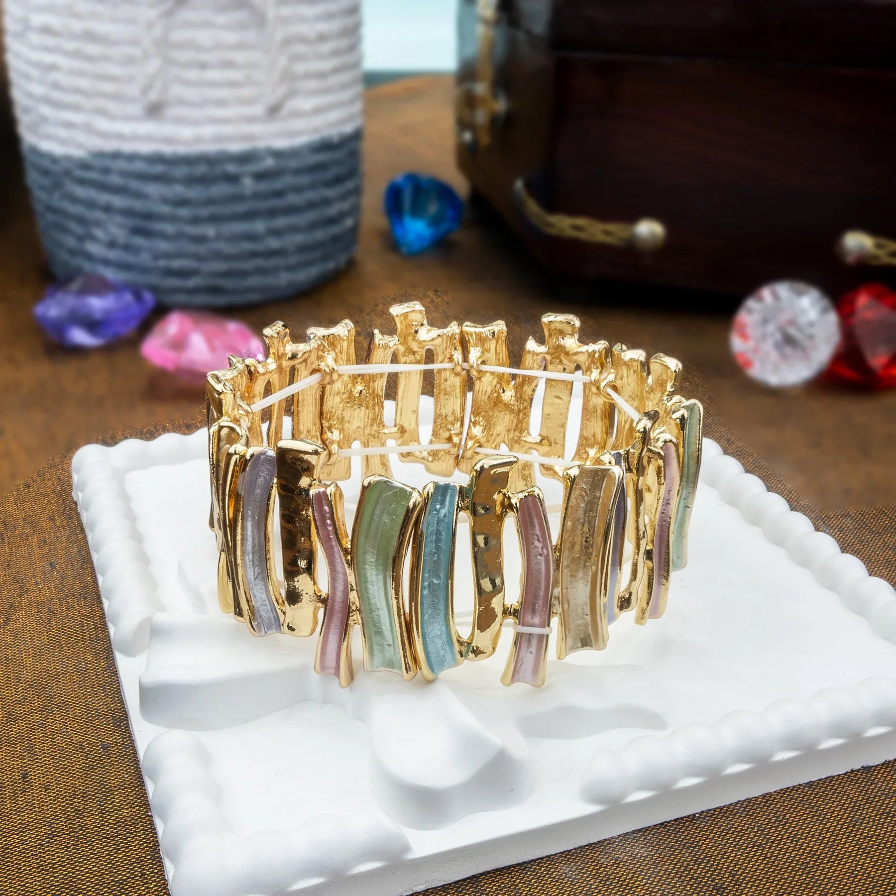 Gold bracelet with colorful stones on a white stand, with gemstones in the background.